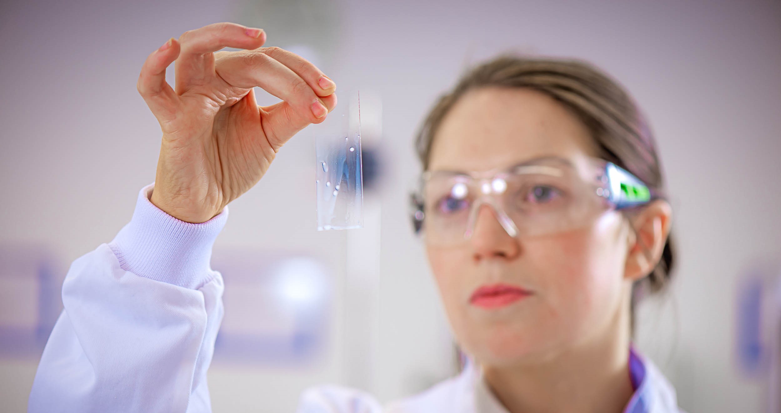 Woman in a lab coat and protective goggles holding a microscope slide.