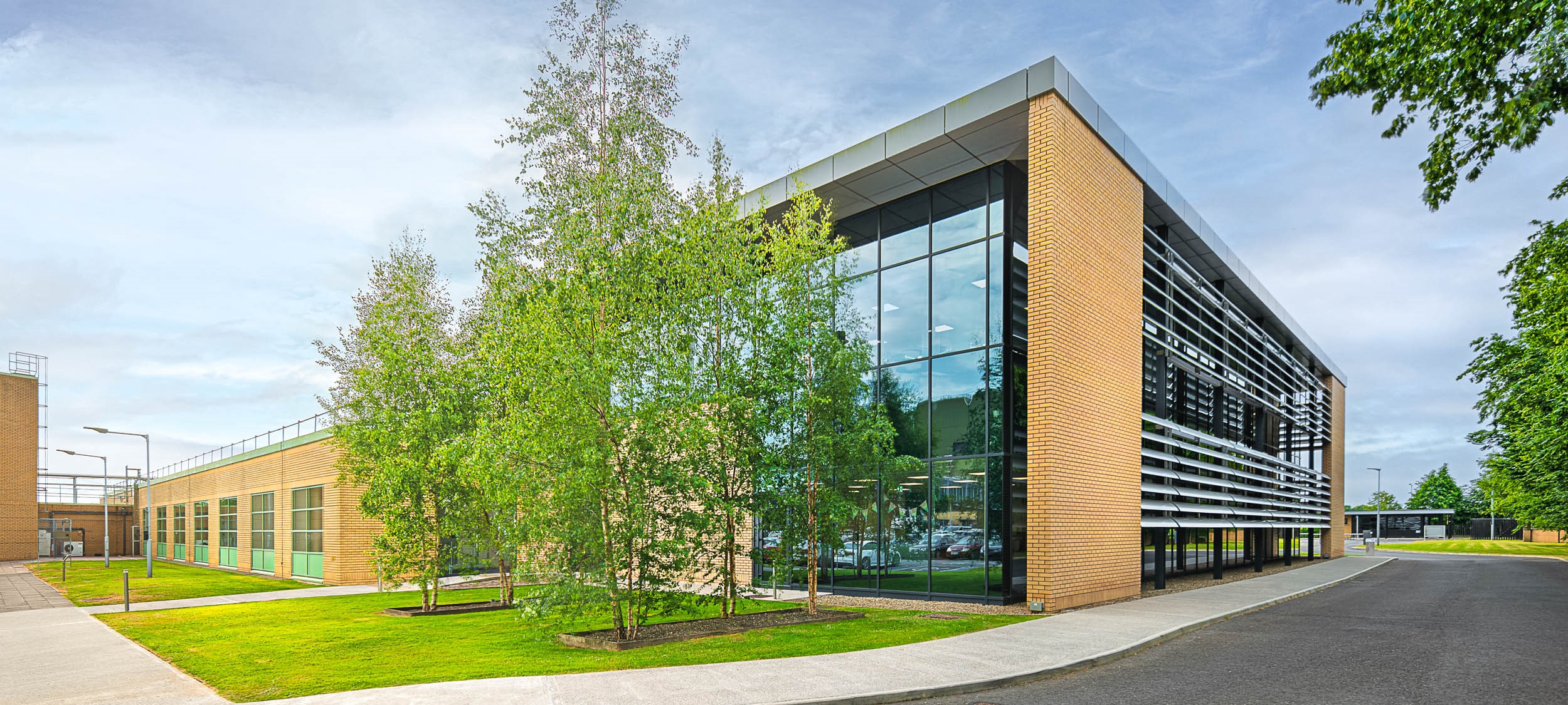 Building with brown brick and floor to wall ceilings, with grass and trees out front, and a blue sky in the background.