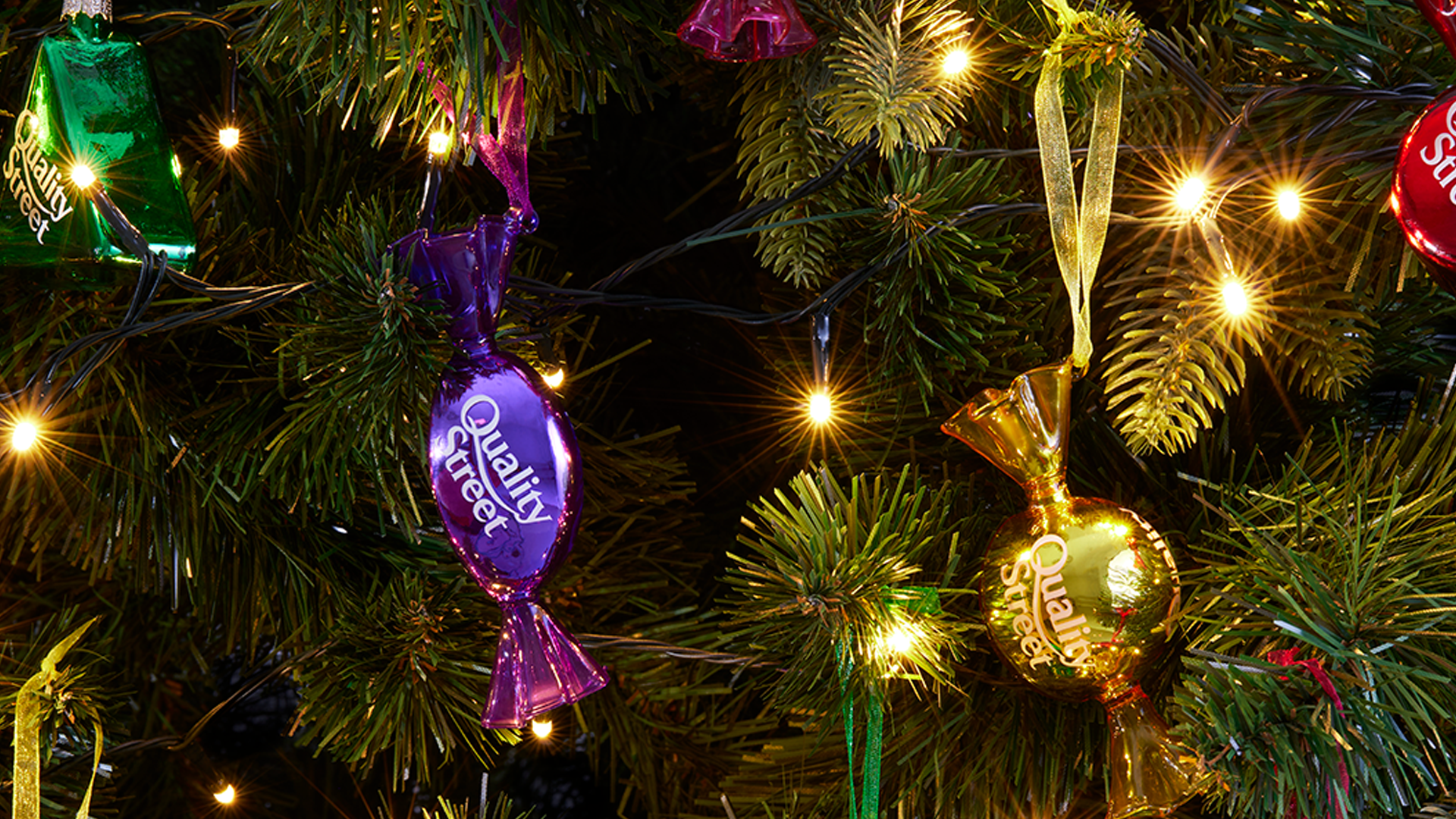A close up photo of two purple and gold Quality Street Christmas baubles, on a green Christmas tree. 