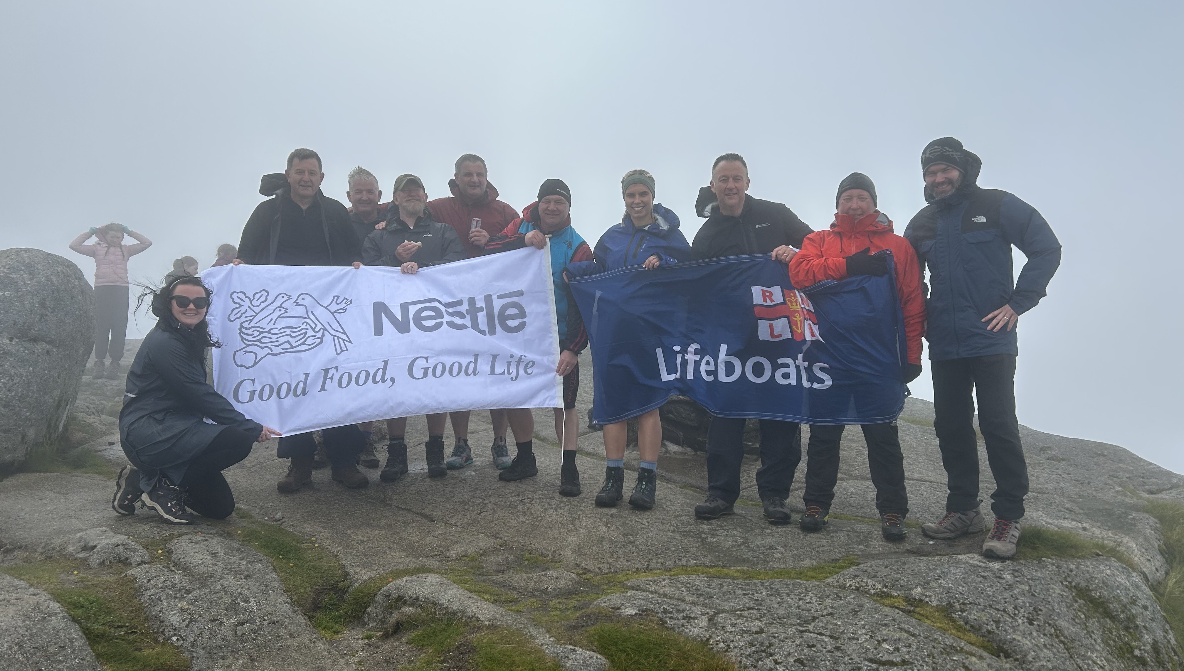 A group of ten people standing on top of a misty mountain with a Nestlé banner and a RNLI banner. 