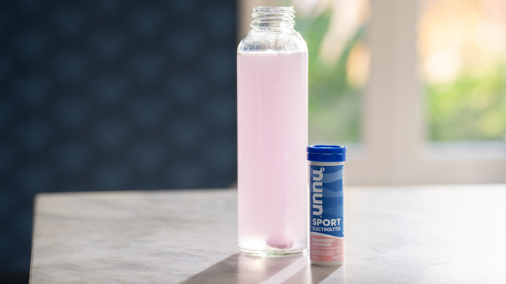 Clear water bottle filled with a light pink liquid next to a small blue container with 'Nuun' written on it on a countertop.