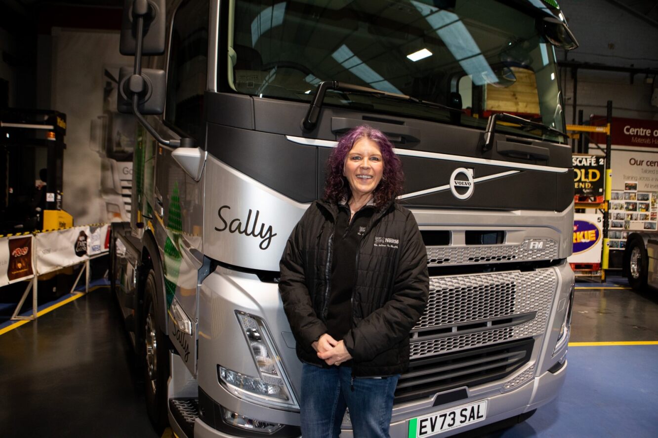 Lady with purple hair standing in front of a silver HGV truck with the name 'Sally' written on the front of the truck. 