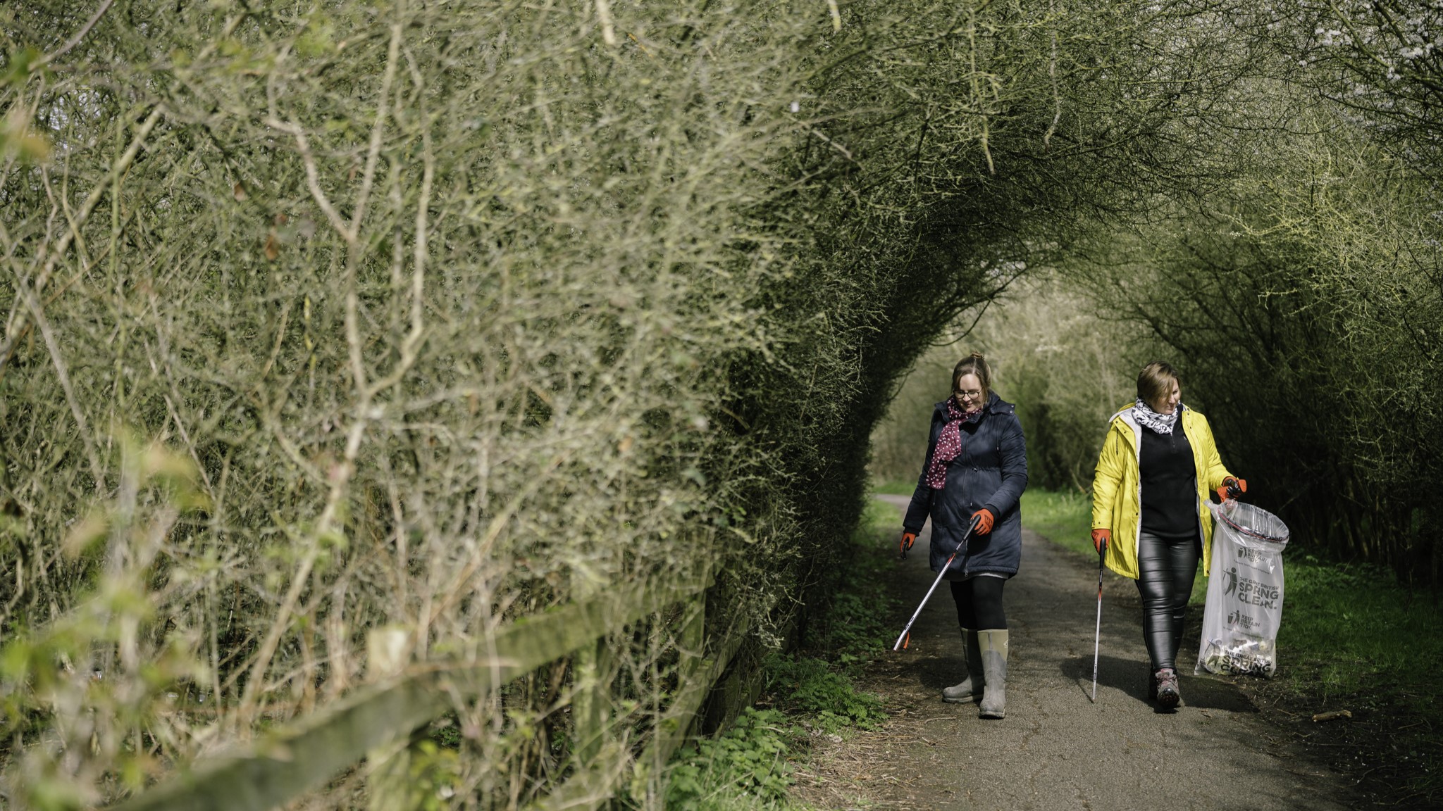 Two women walking through an arch in a forested area holding bin bags and litter pickers. 