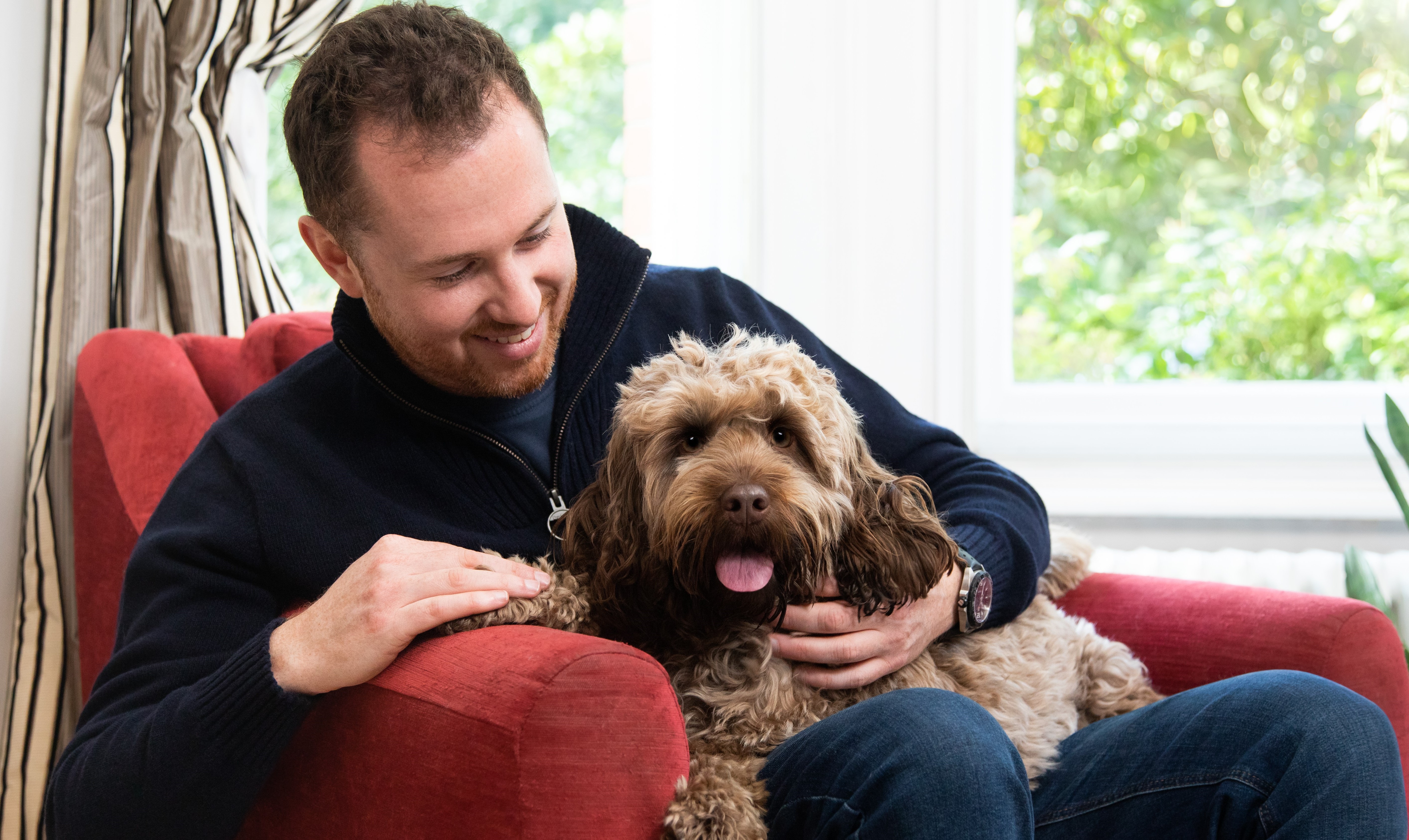 A white male sat on a red chair with his dog on his lap.
