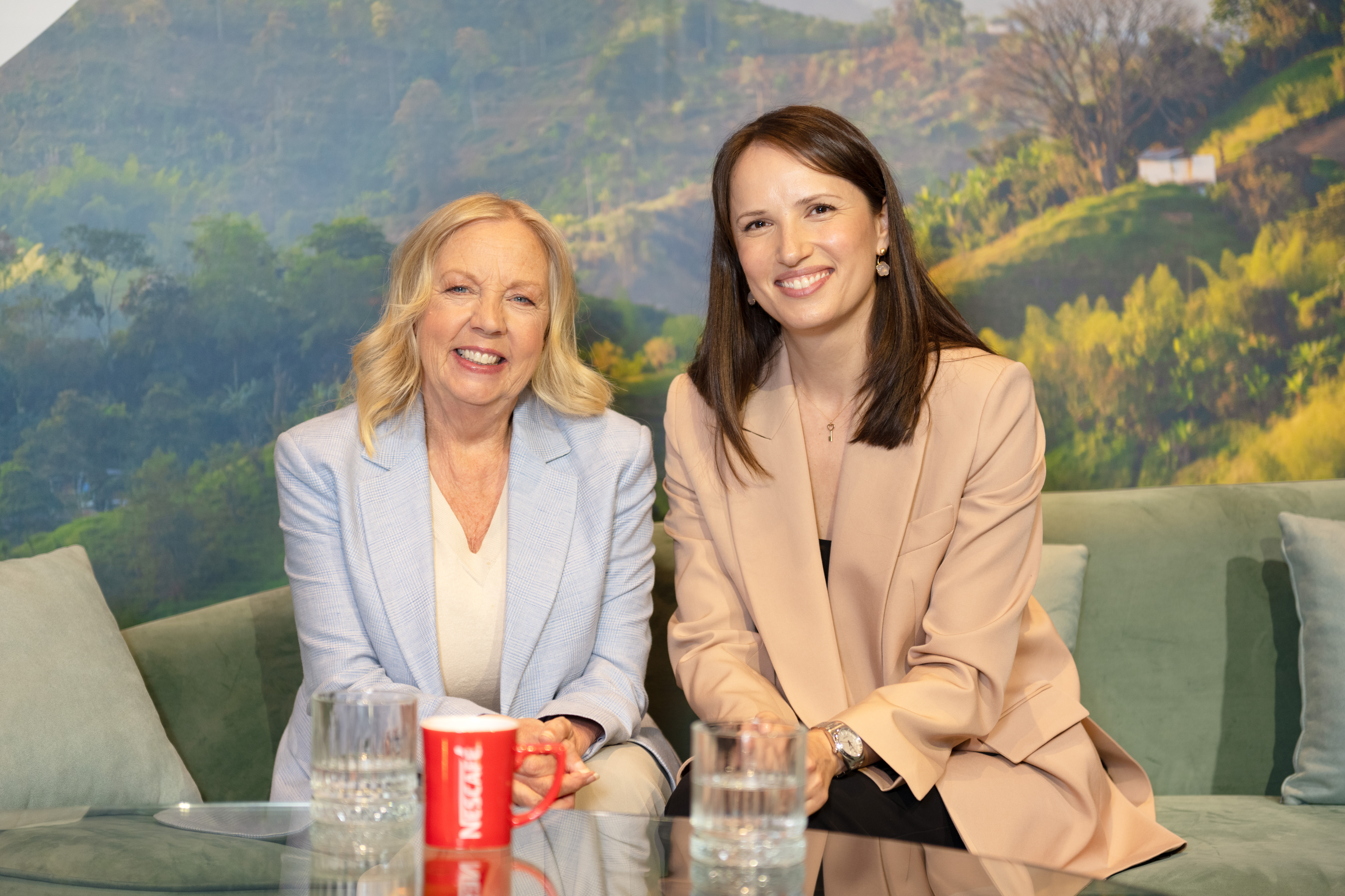 An image of two white females. Deborah Meaden (left) and Katya Simmons (right) sat on a sage green seat, smiling together.