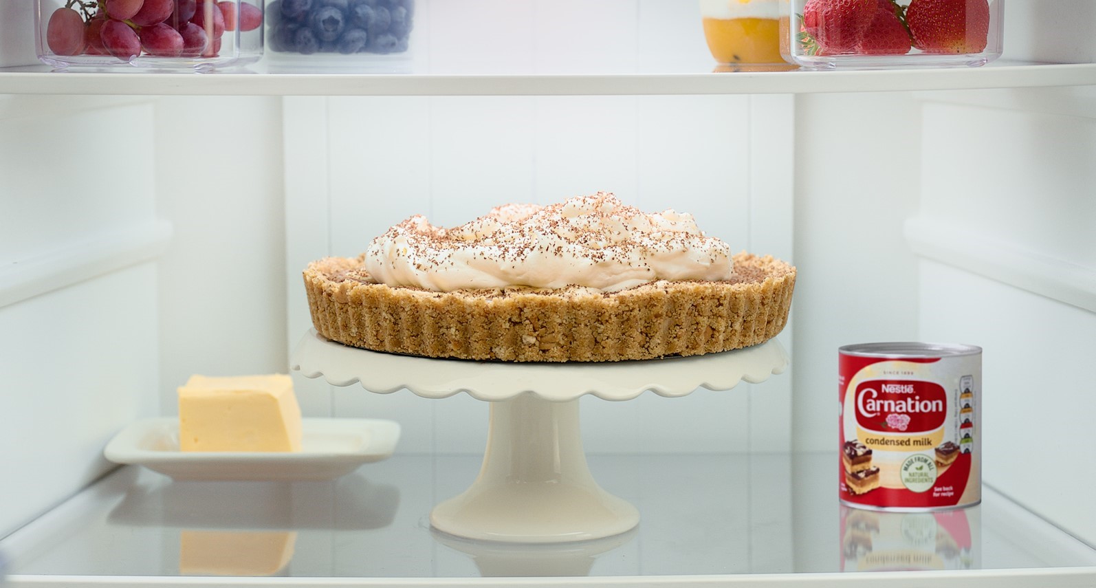 A landscape still image of a fridge with a banoffee pie with cream next to a red and white tin of Carnation.