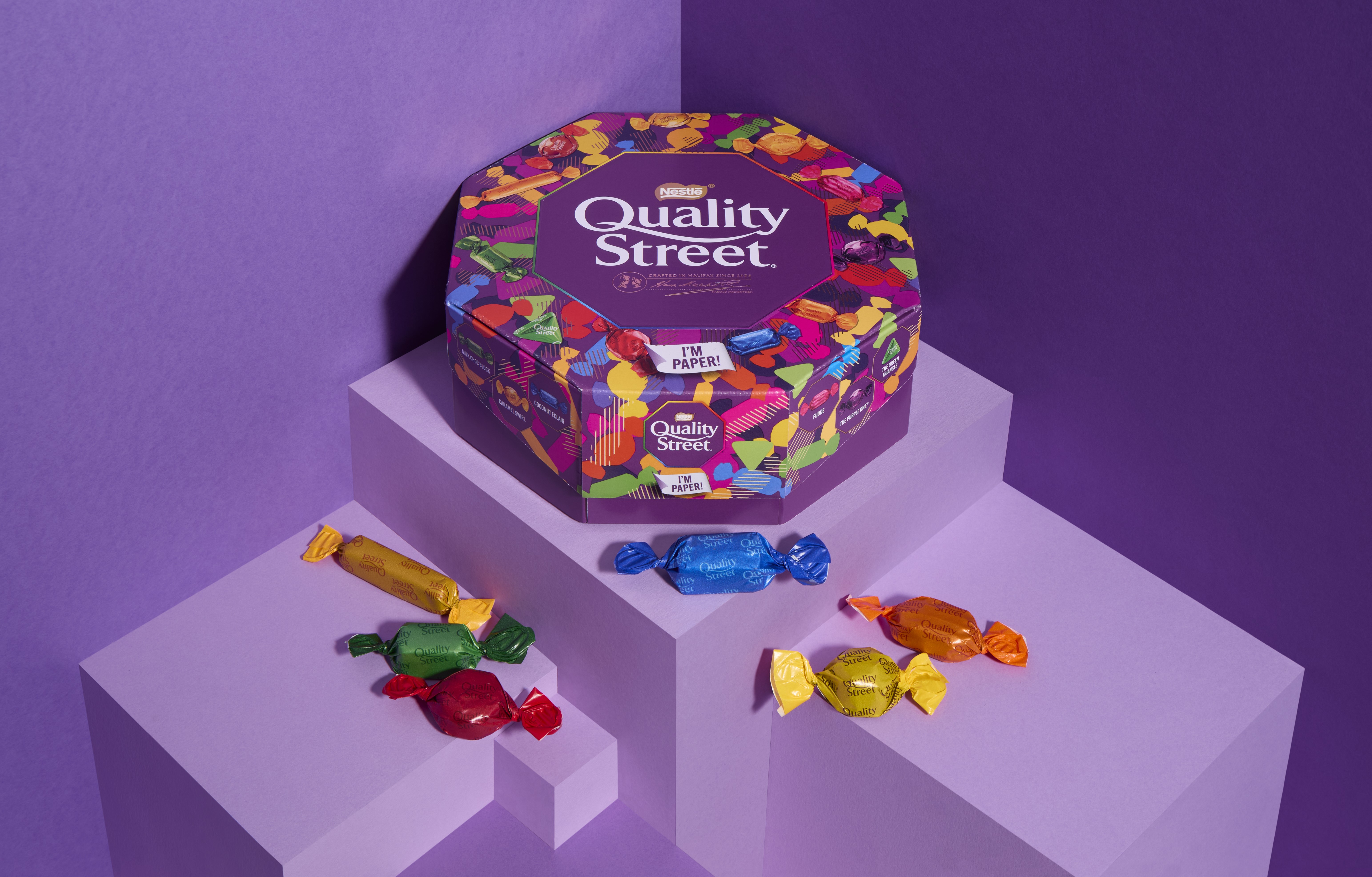 An image of a dark purple Quality Street paper tub on a light purple step with a selection of multicoloured Quality Street treats around it. 