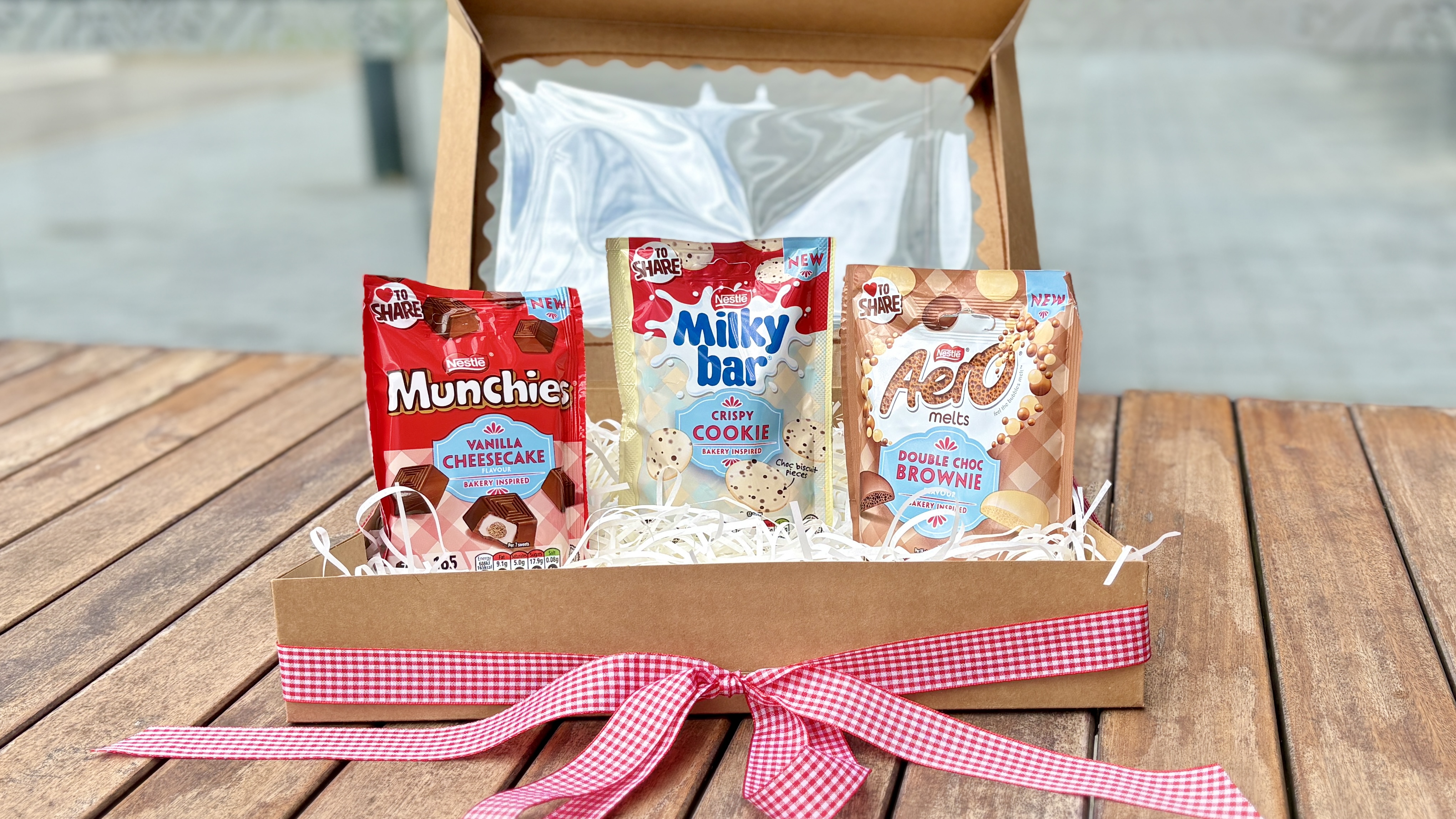 A landscape image of a a rustic-style box with a red gingham bow on, filled with Munchies, Milkybar and Aero sharing bags.
