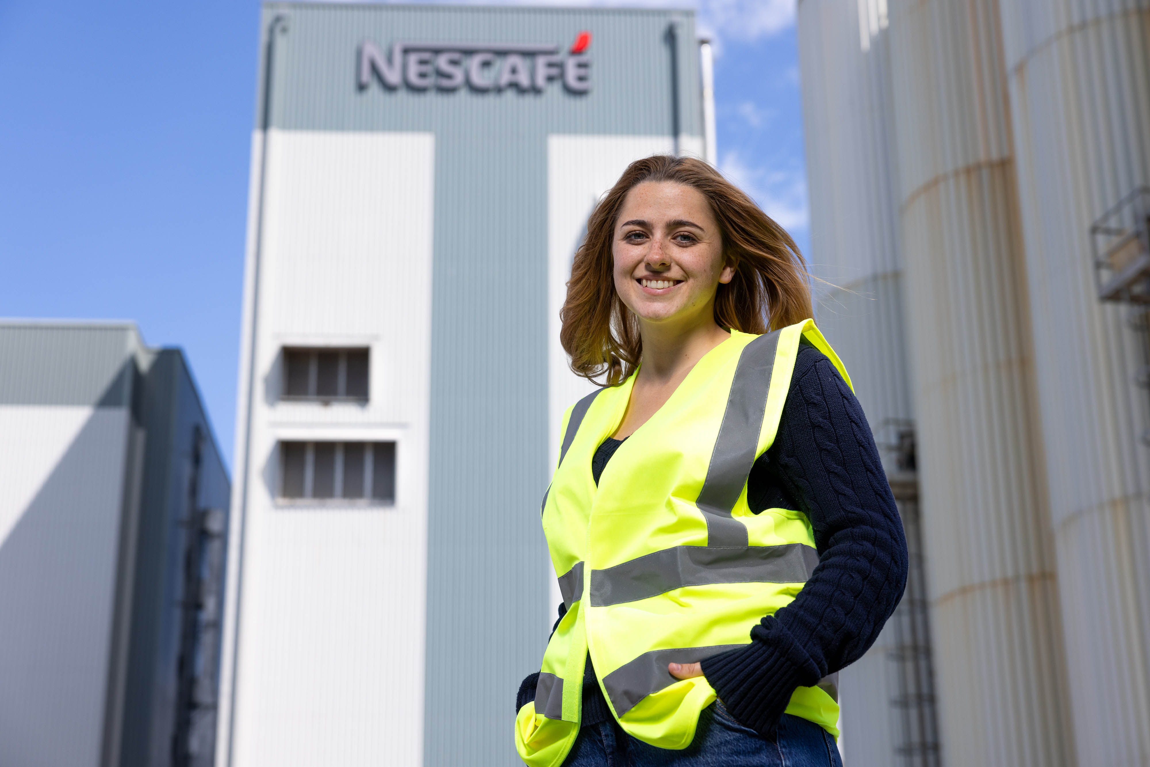 A landscape image of a white female with long brown hair, smiling, in a high-vis jacket out side a Nescafé factory. 