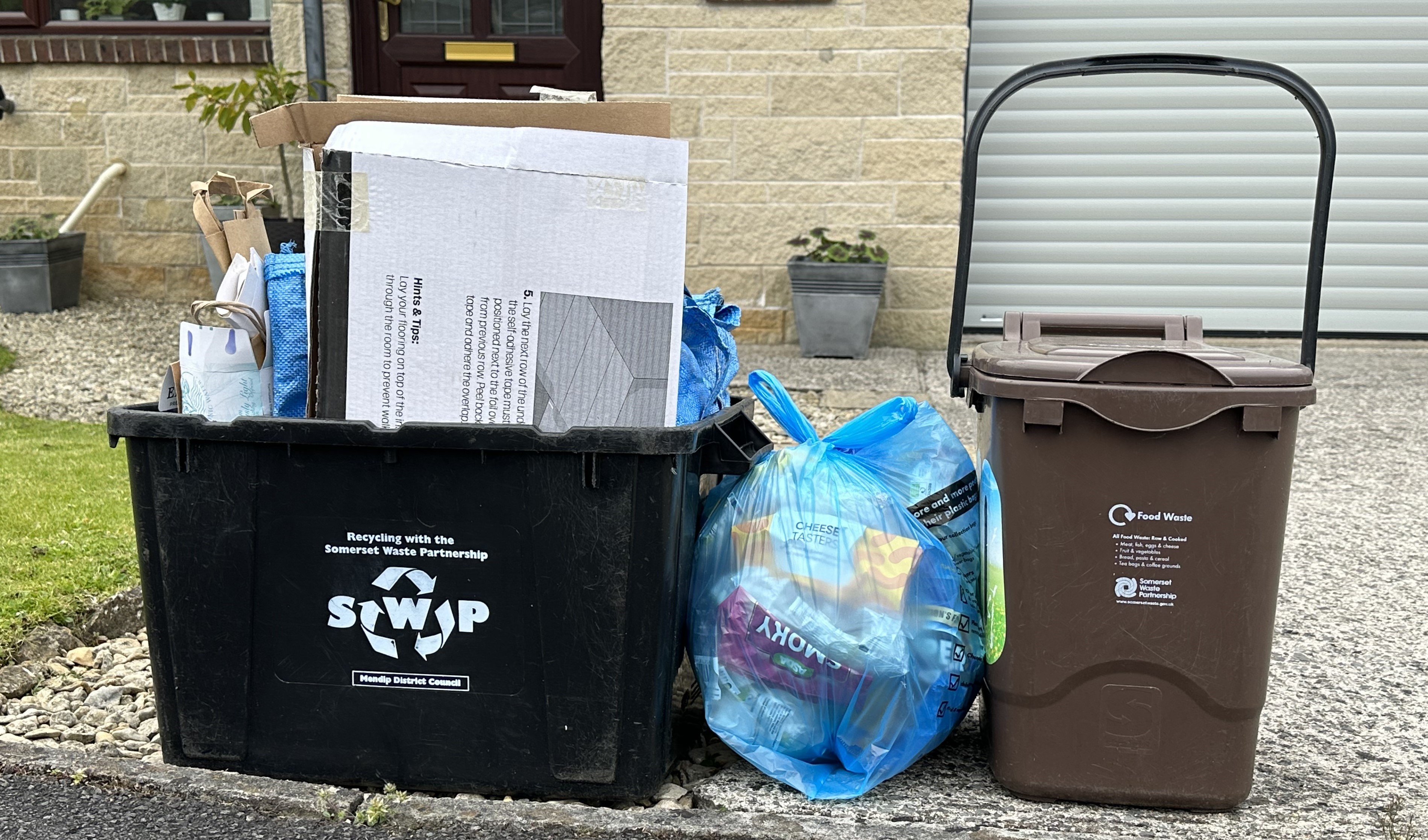 A landscape image of a blue plastic recycling bag, next for a black bin box and a brown compost bin, on a curb side. 
