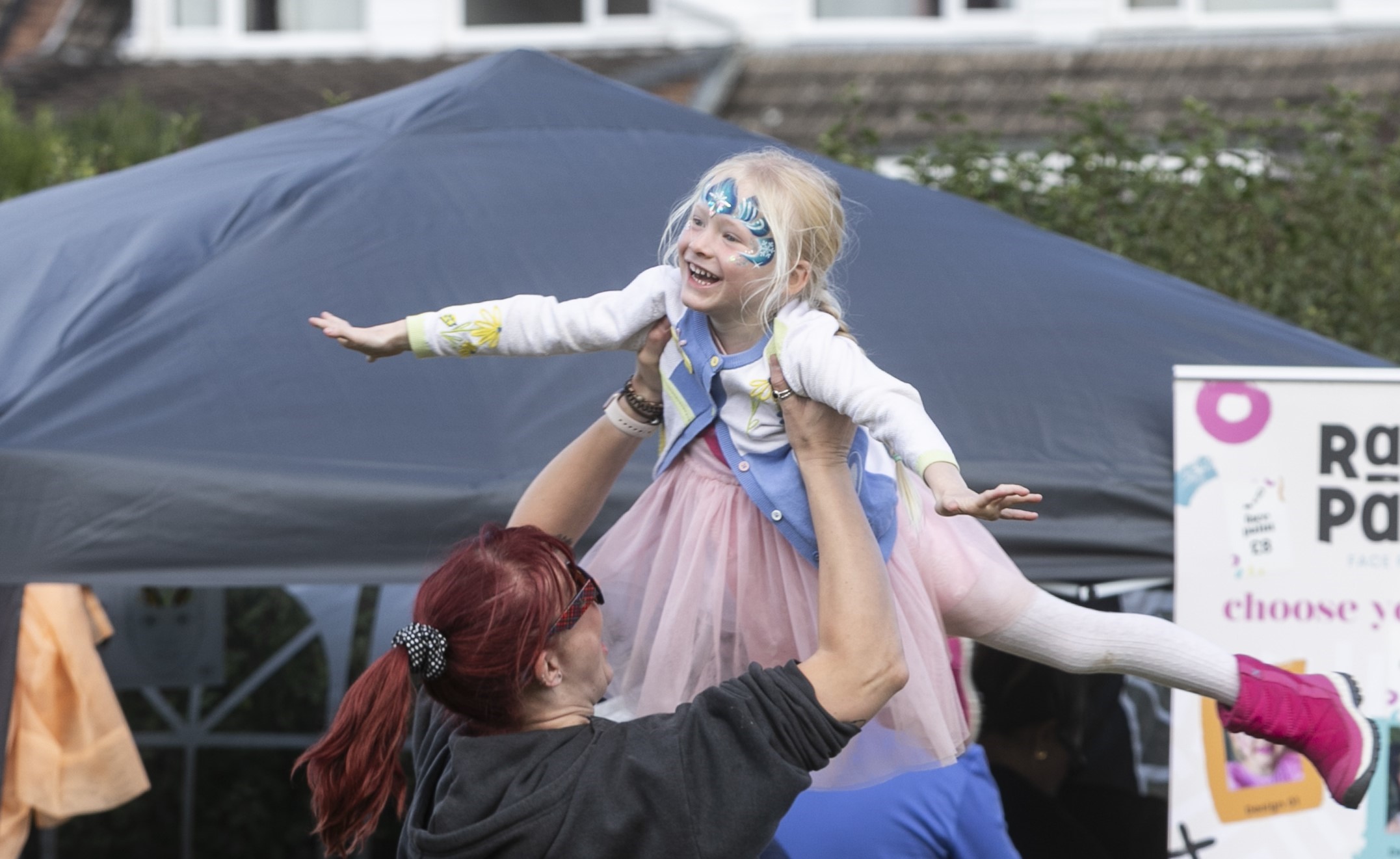 An image of a child smiling with butterfly face paint on, being held by her mother. 
