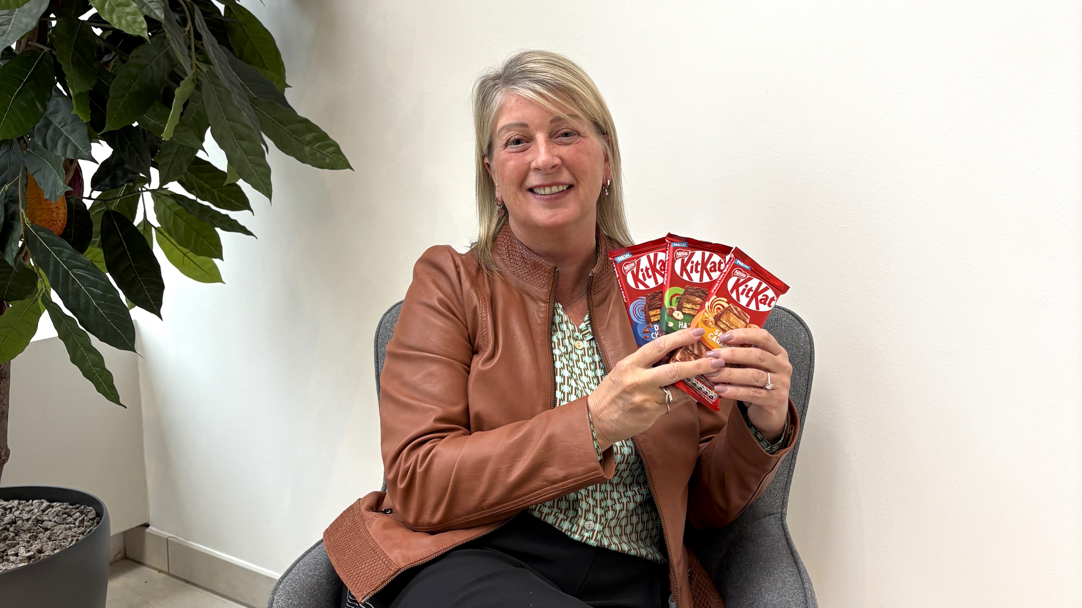 A lady sitting on a chair and smiling at the camera, whilst holding three KItKat sharing bars.