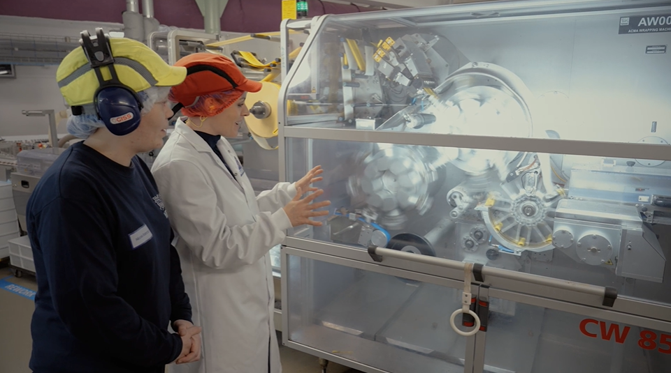 A landscape image of two women in neon hardhats and factory PPE, looking at a large silver paper sorting machine with cogs. 