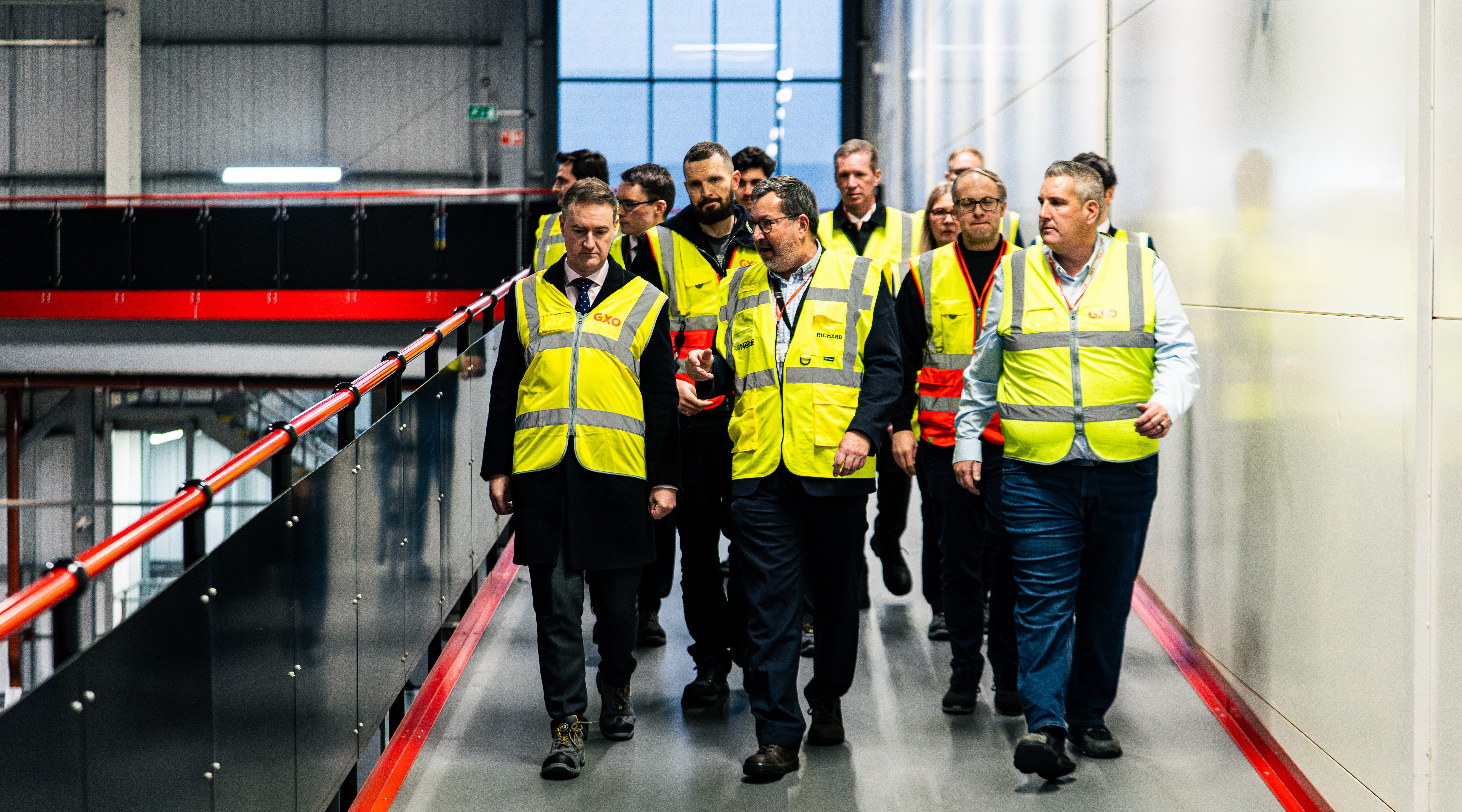 A landscape image of a group of males in yellow hi-visibility jackets walking down the walkway of a distribution centre.  