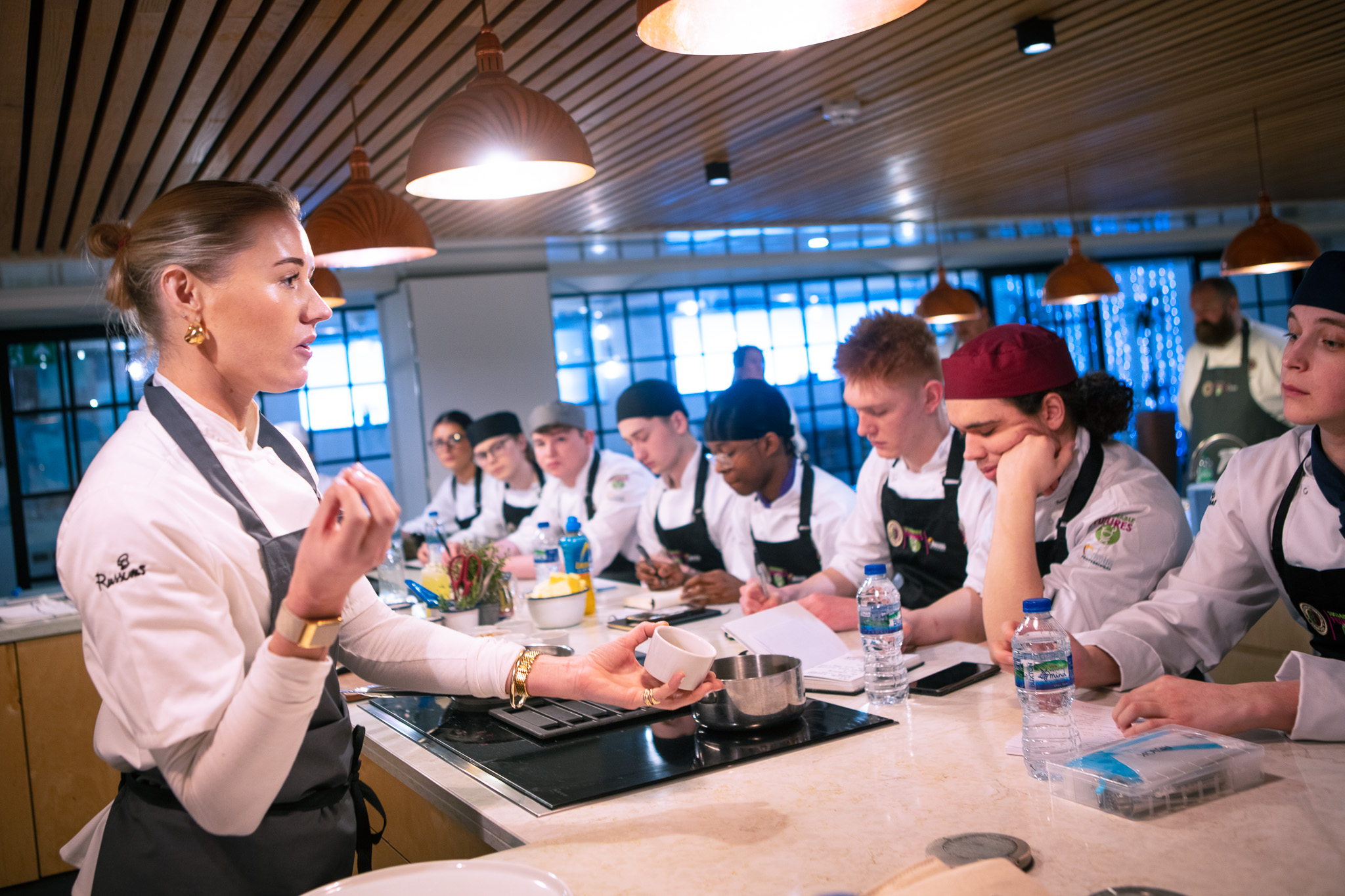 A landscape image of a female chef instructing a mixed gender group of young chefs in a working kitchen.