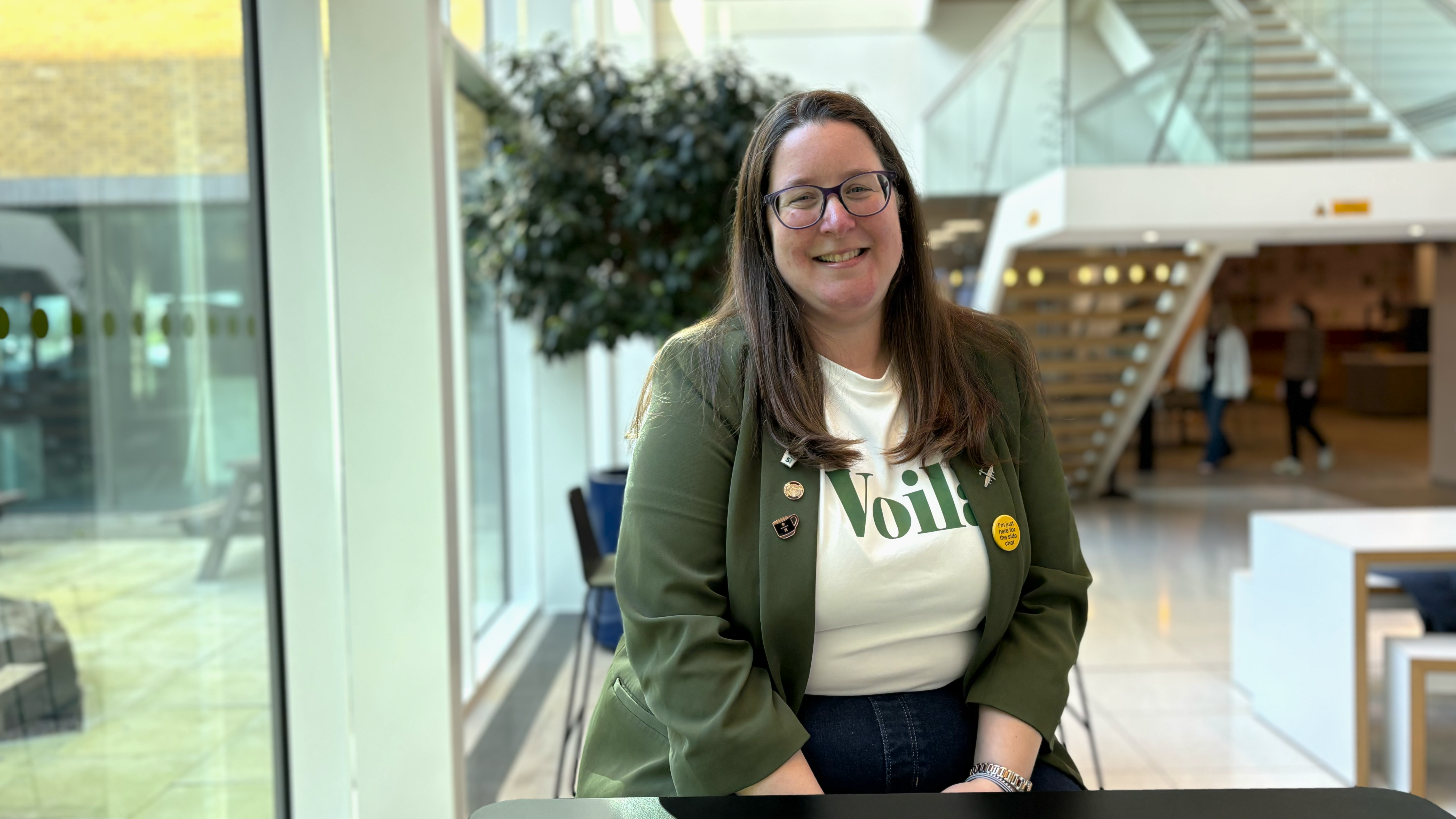 Woman sitting in an office environment wearing a white top and a green blazer.