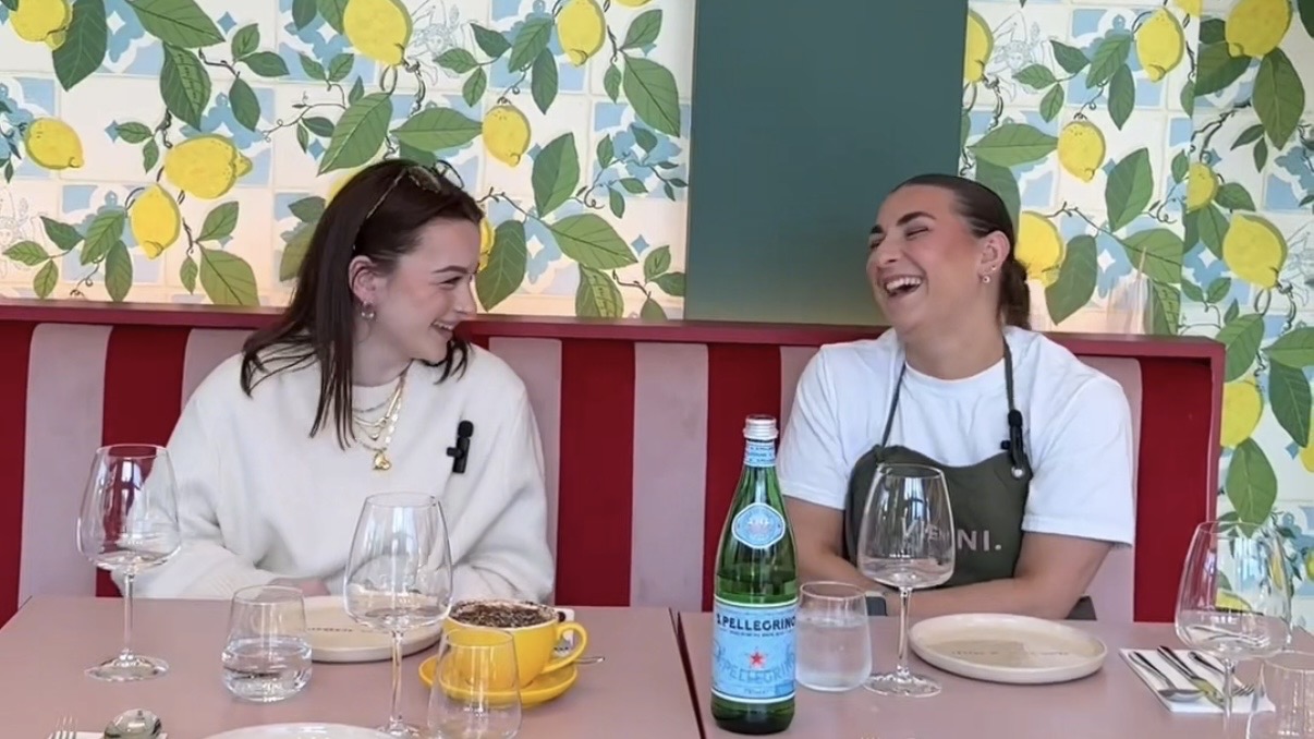 A landscape image of two white women with brown hair, one is a chef in an apron, sat at a dining table in a colourful room. 
