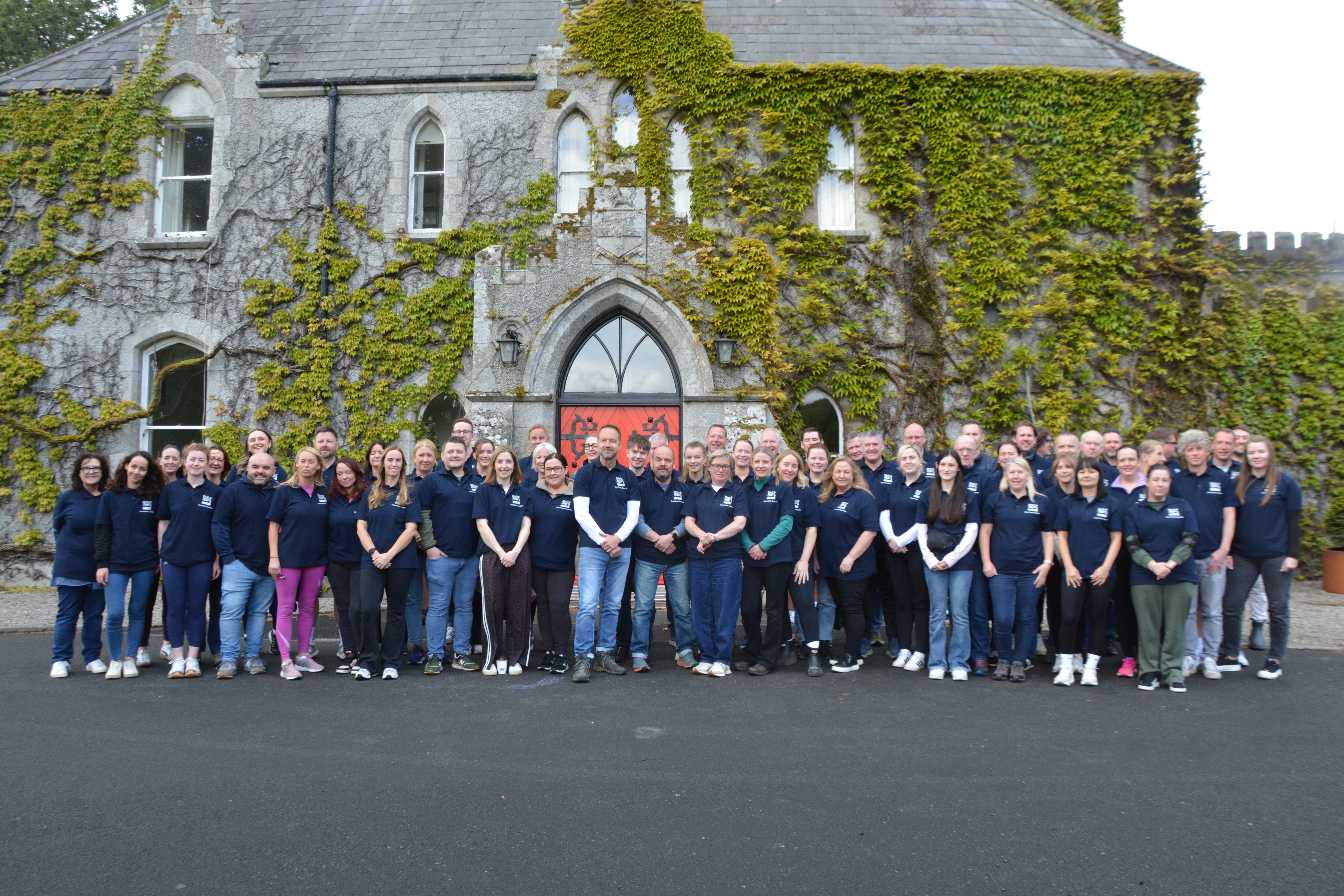 Group of colleagues all standing together in front of a building covered with ivy, Barretstown location.