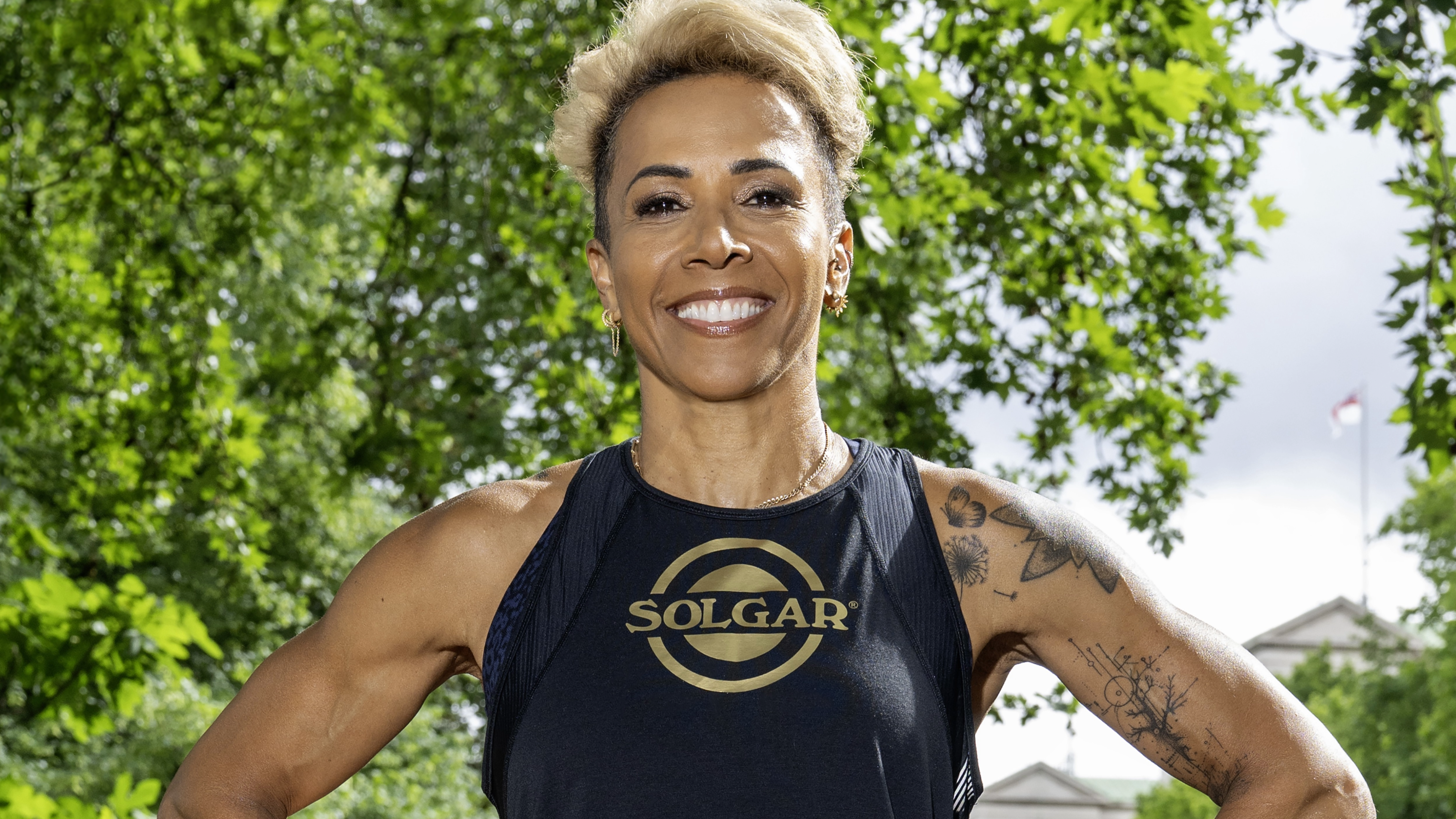 A landscape image of Dame Kelly Holmes with her arms at her side, smiling, in a black vest top. 