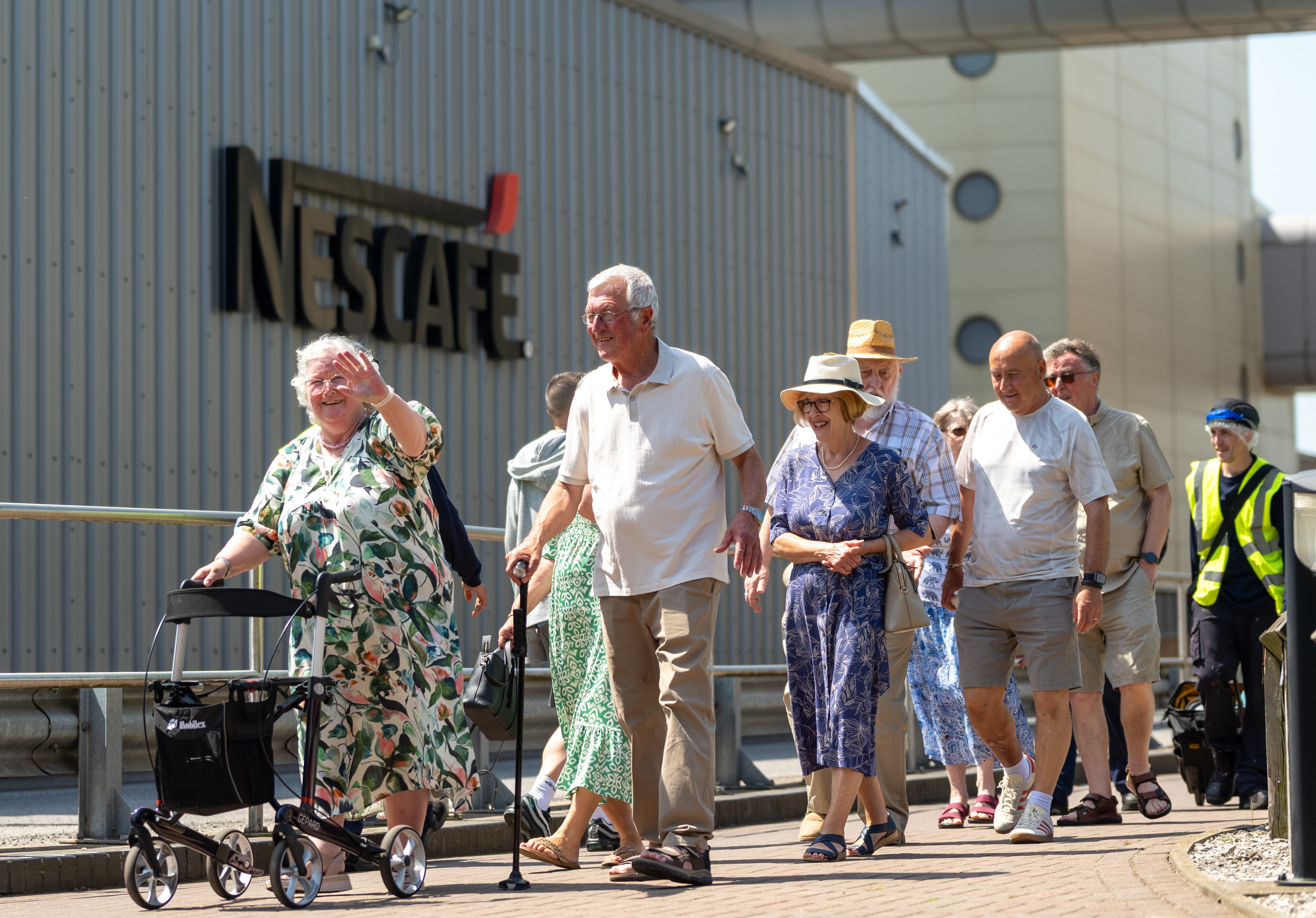 A landscape image of a mixed gender group of local pensioners, smiling and waving outside a Nescafé factory. 