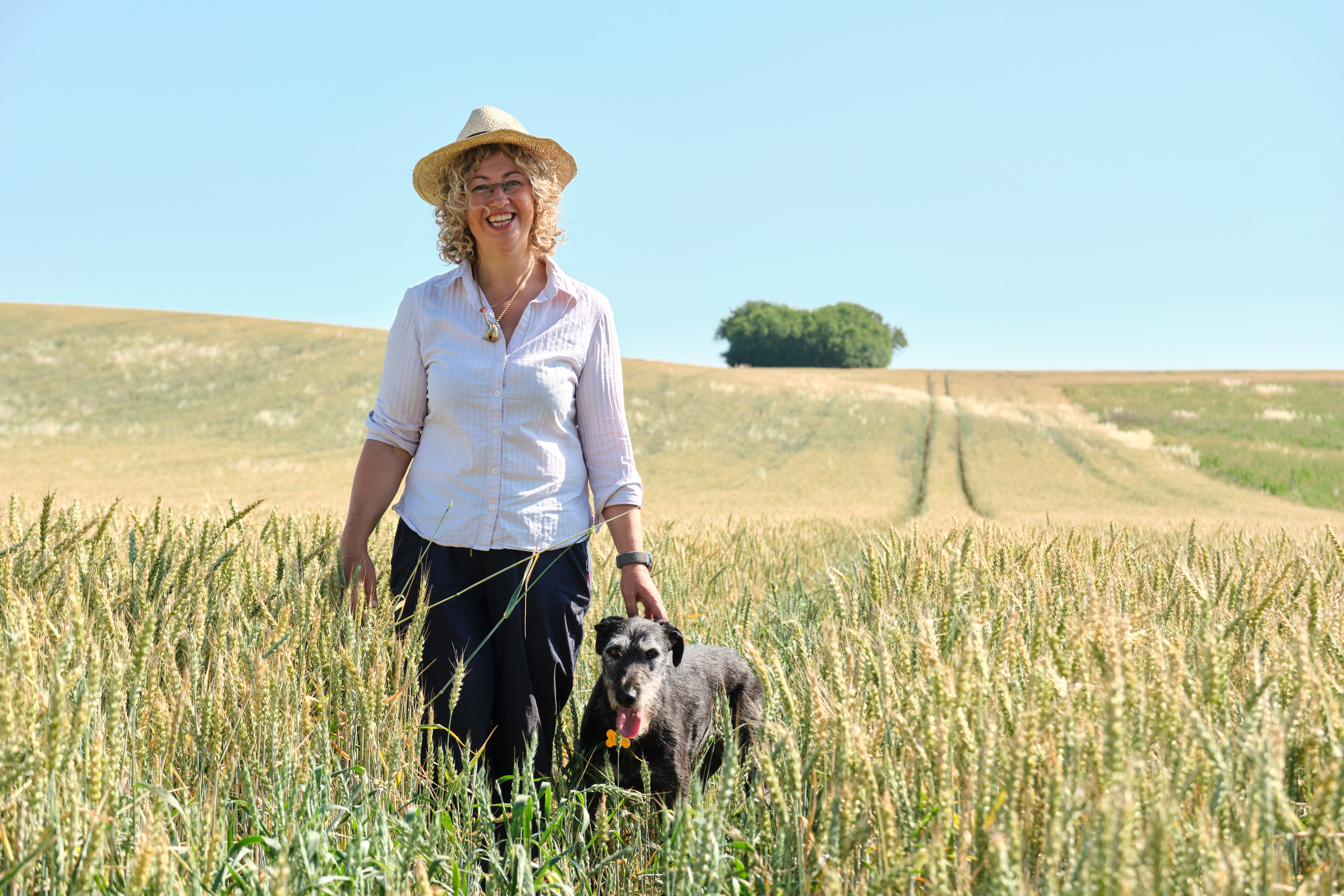 A farmer standing in a wheat field with her grey dog by her side.