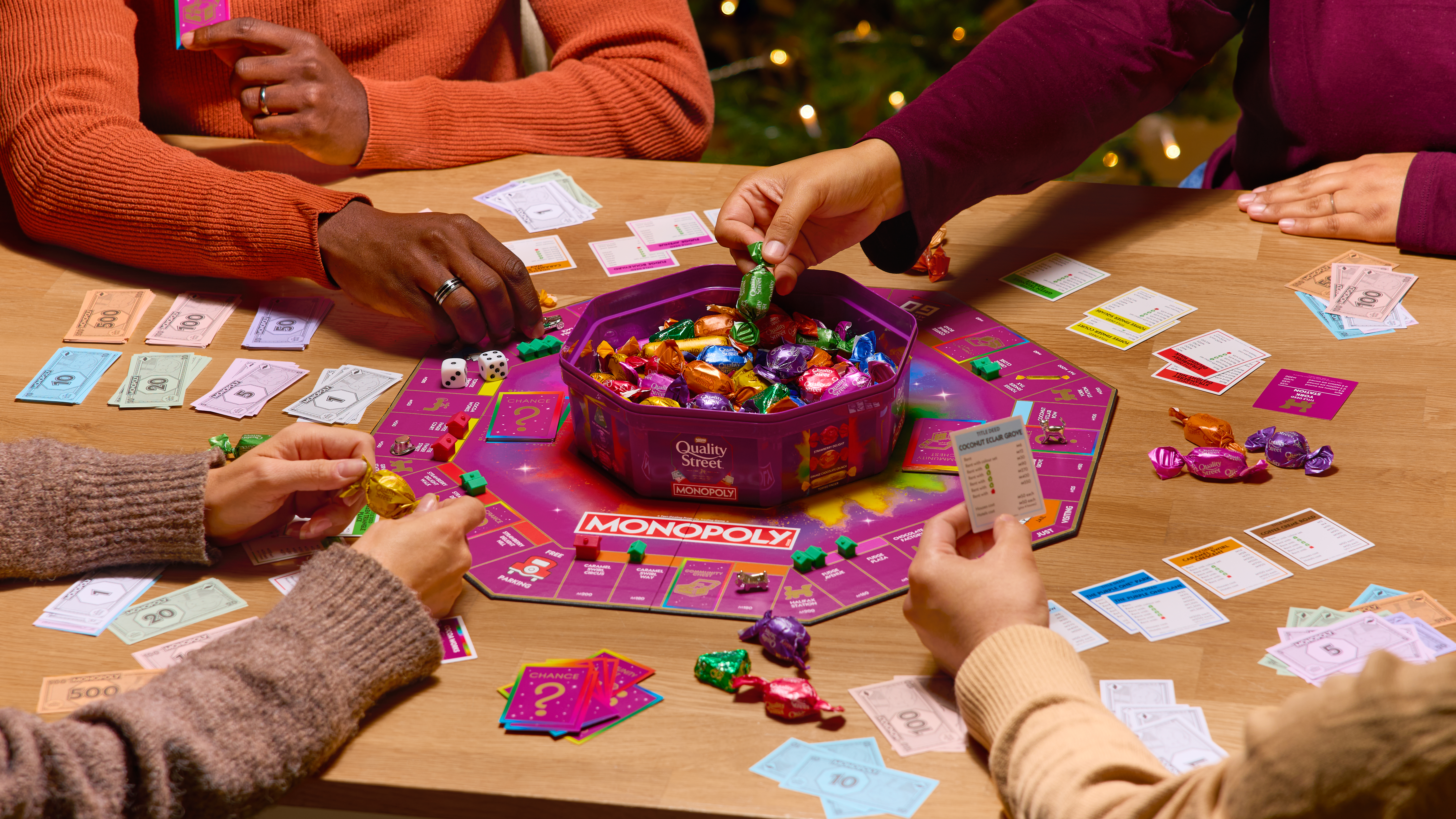 A group of people playing Monopoly with a Quality Street tub in the middle