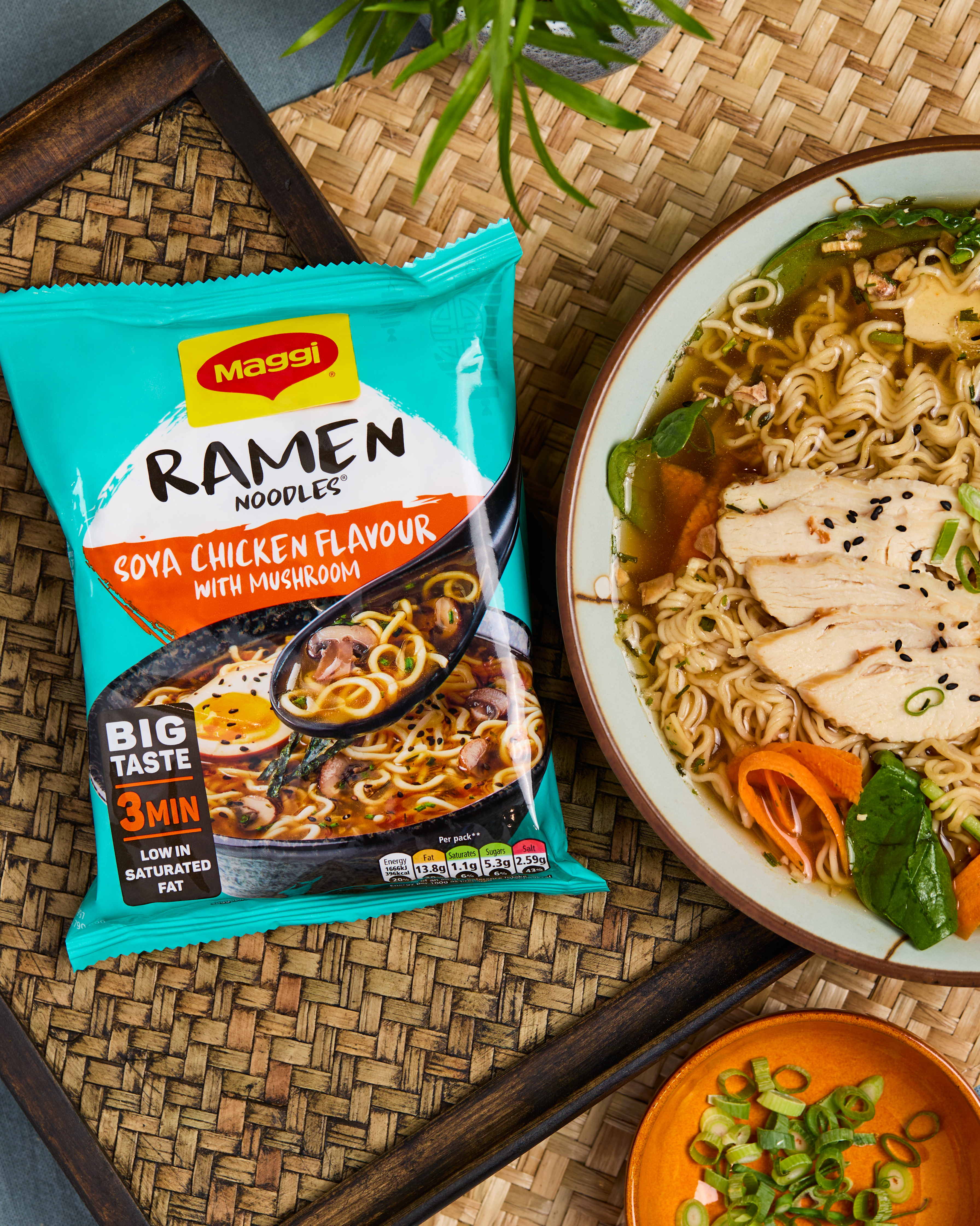 Packet of Chicken Soya Flavour Maggi Ramen Noodles next to a bowl of noodles and various ingredients, on a wooden placemat.