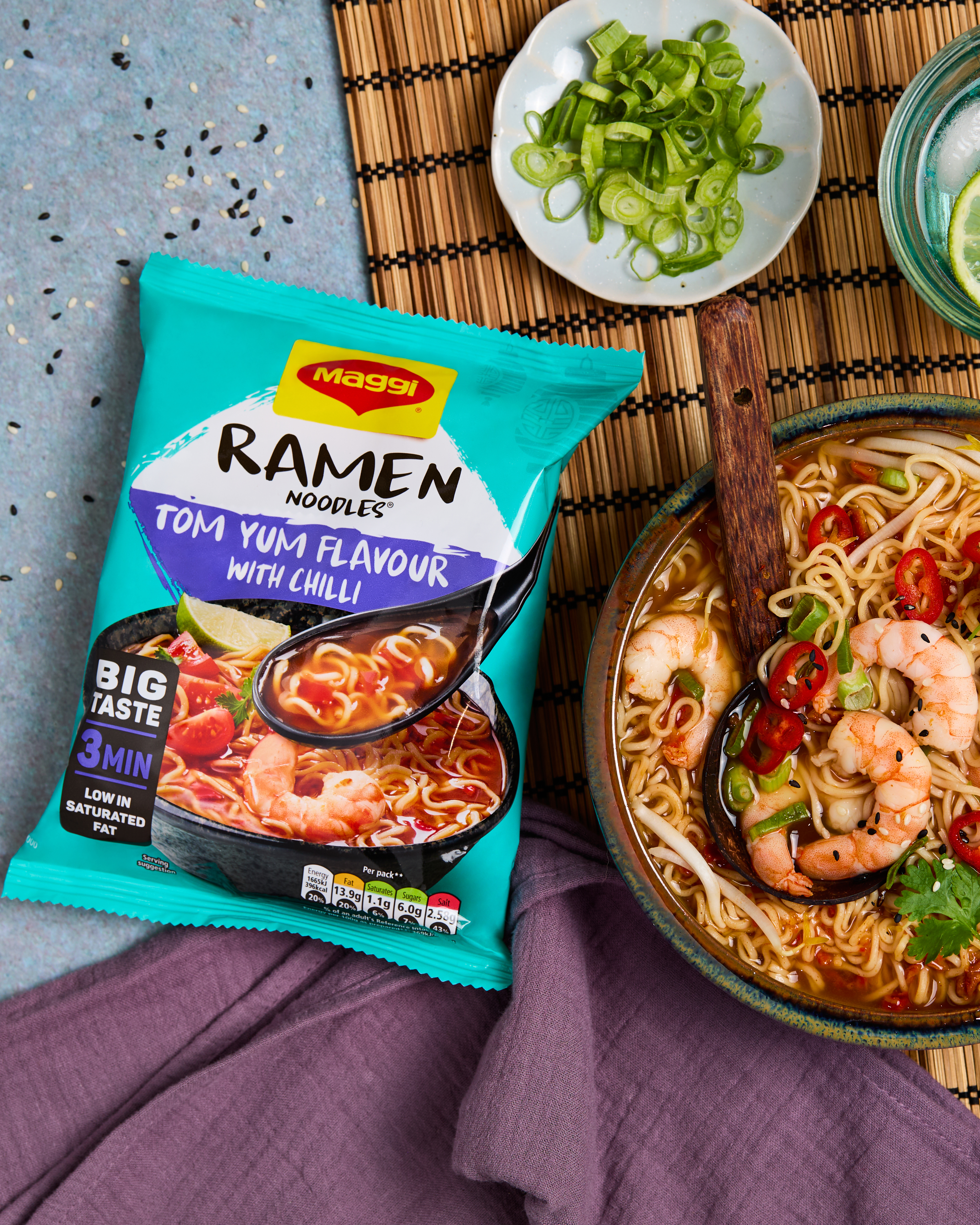 Packet of Tum Yum Maggi Ramen Noodles next to a bowl of noodles and prawns sitting on a wooden placemat with purple napkin.