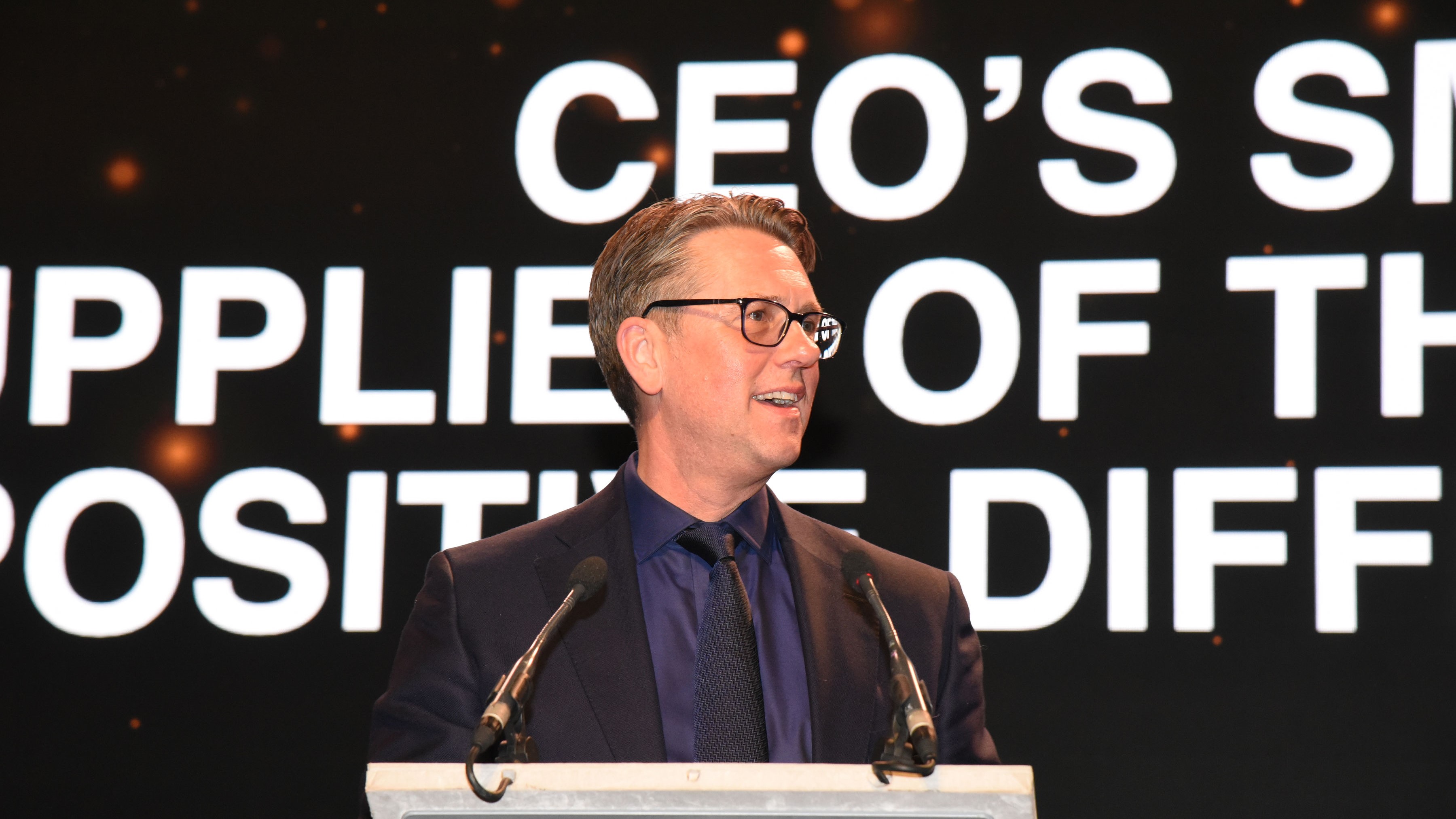 Man standing in front of a podium, in the back a screen reads CEO's Supplier of the Year, Positive Difference Award