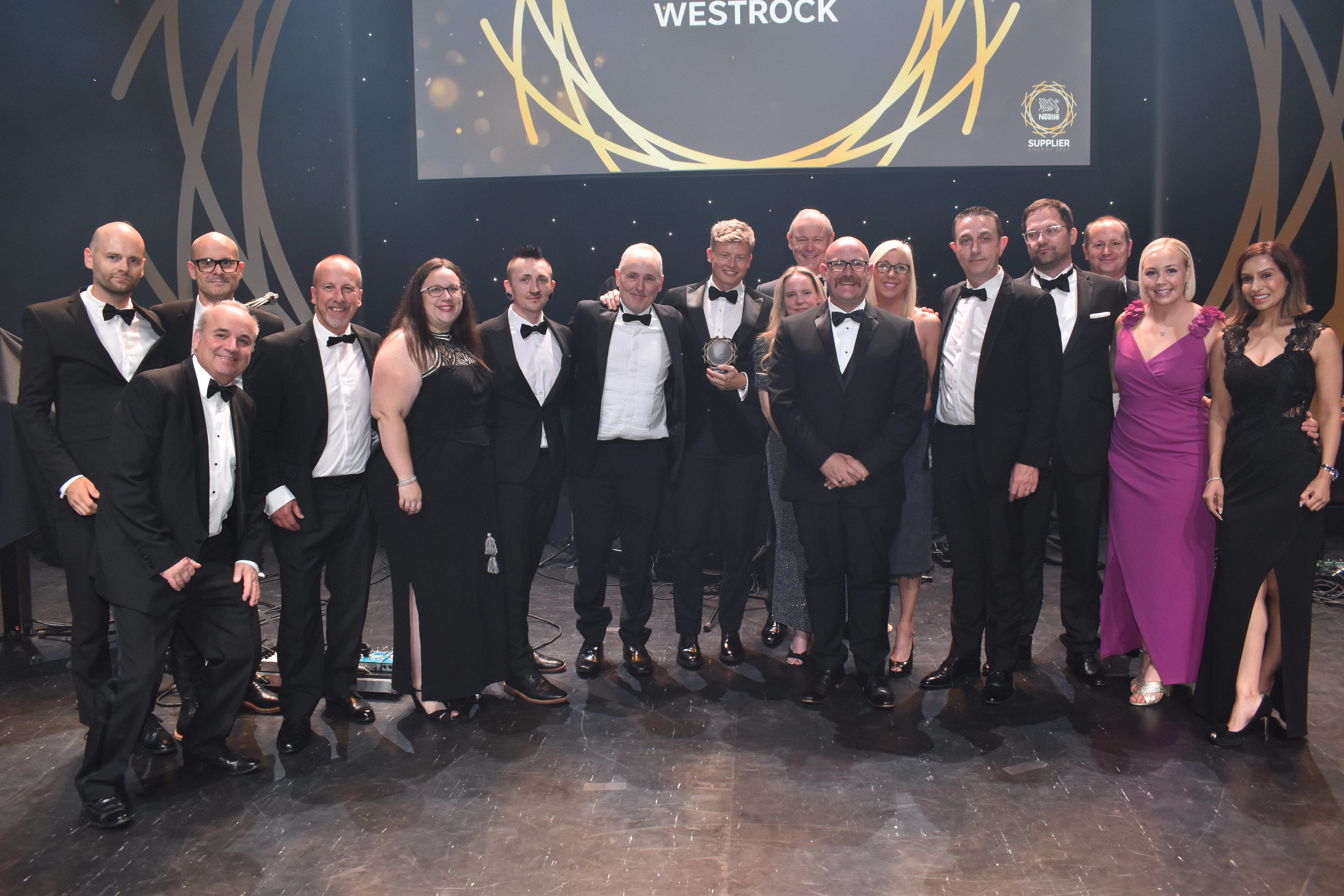 Sixteen people on stage and surrounding an award.