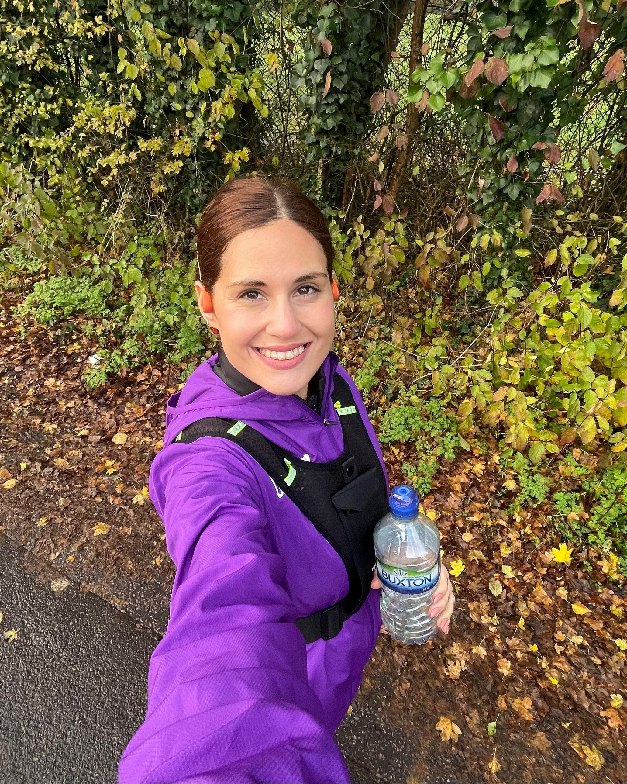 Young woman in athletic clothing holding a Buxton water bottle whilst outside amongst trees.