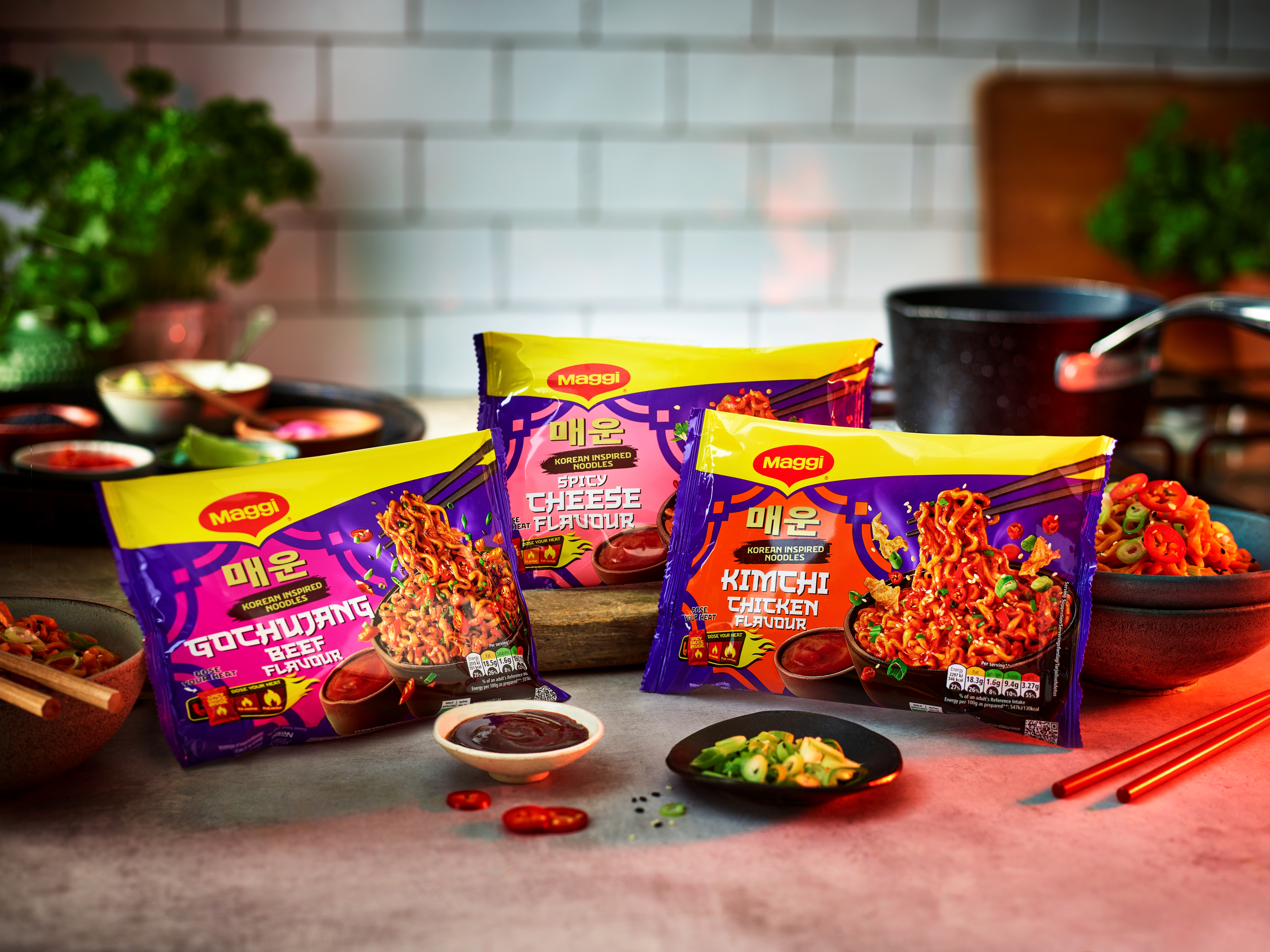 Three packets of the new Maggi Korean-inspired noodles on a kitchen countertop beside ingredients and utensils.