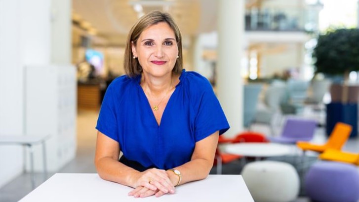 Woman wearing a blue outfit, sitting at a table and looking into the camera.