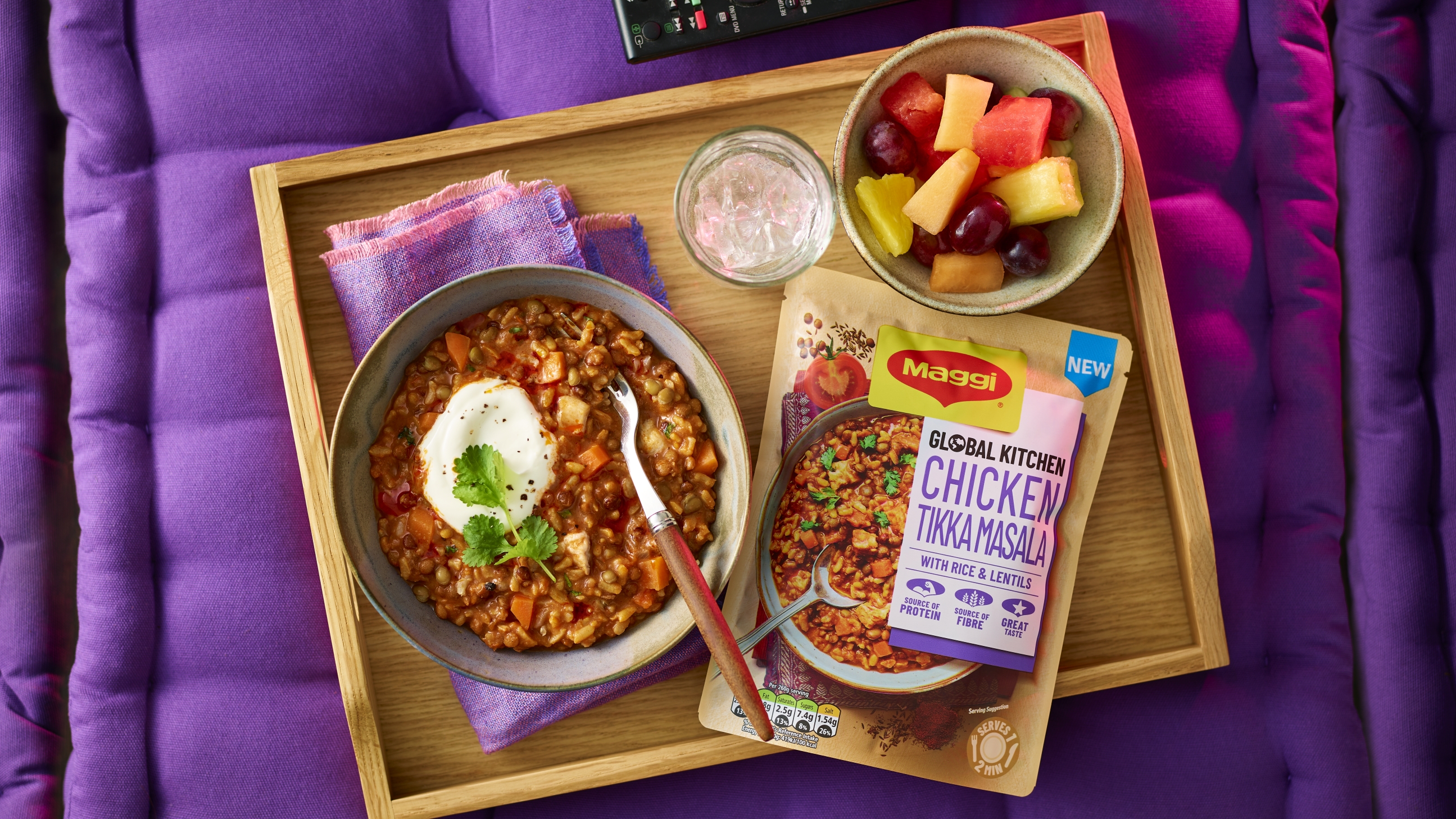 Bowl of Chicken Tikka Masala sitting on a tray next to a ready meal pouch, the tray is sitting on purple bedding.