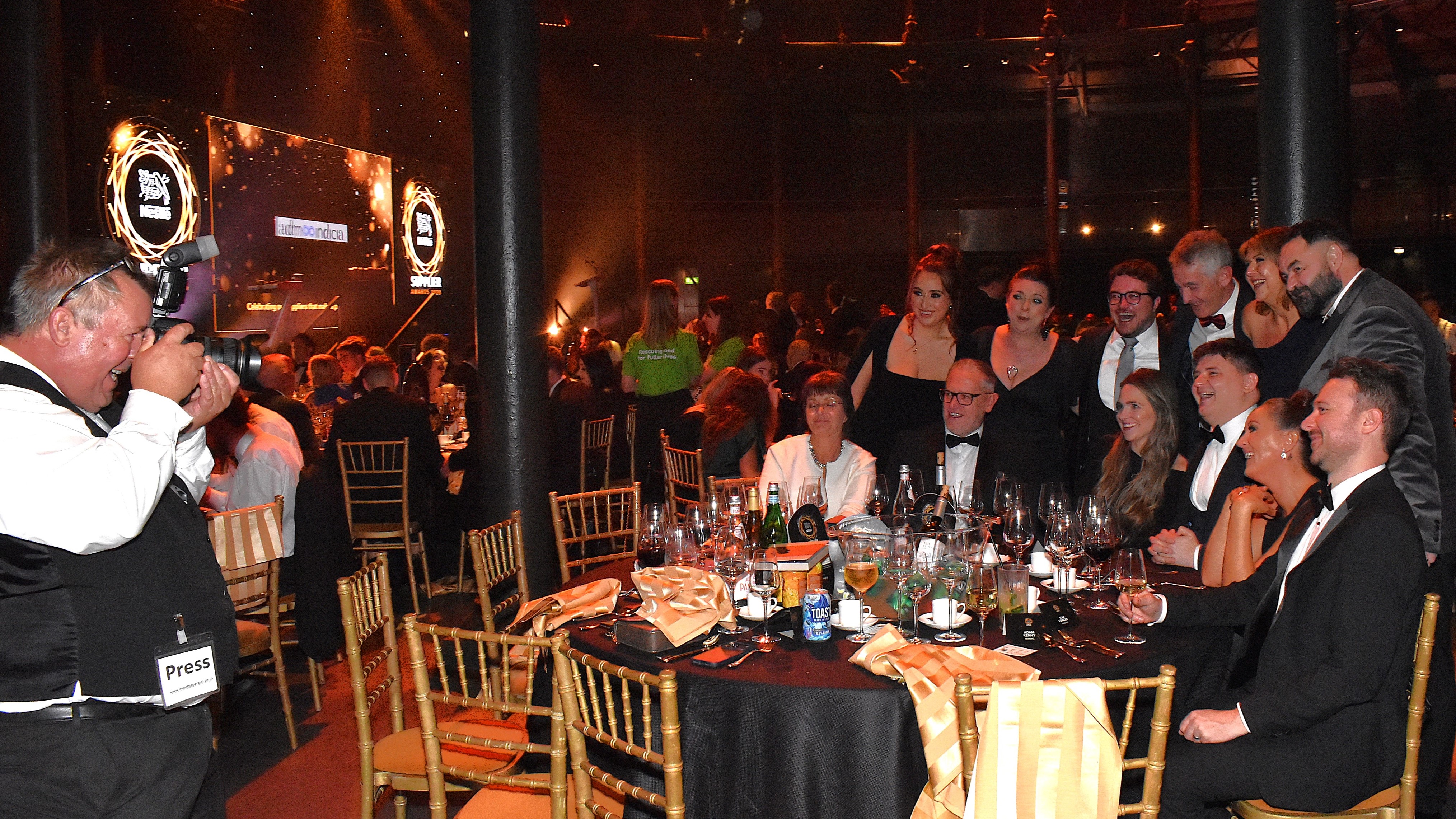 A photographer taking a picture of a table of around ten guests gathered together smiling.