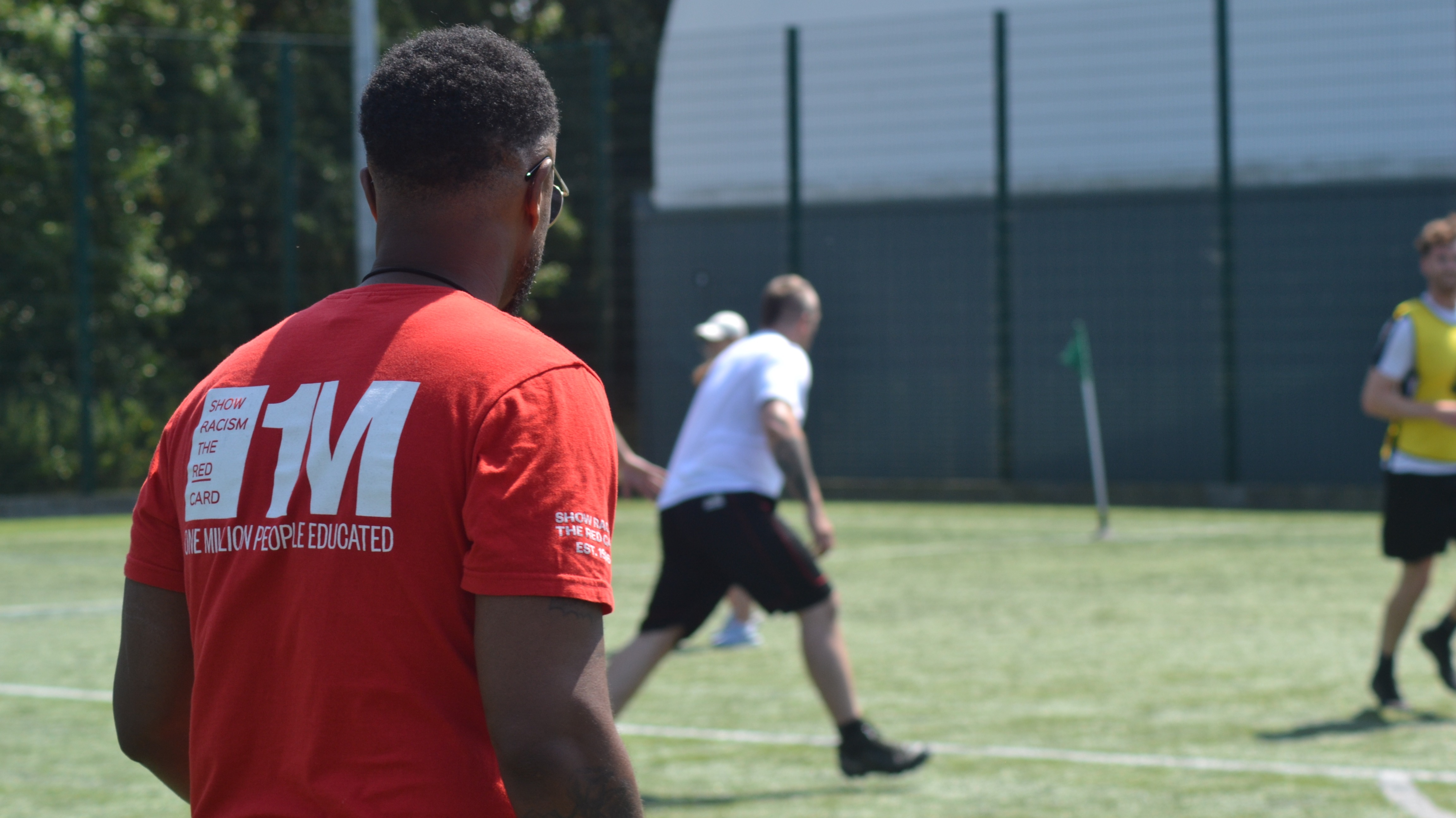 Back of someone wearing a Show Racism the Red Card t-shirt whilst they stand on a football pitch with players in the back.