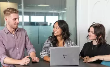 Three people seated at a meeting table discussing work while using a laptop in an office environment.