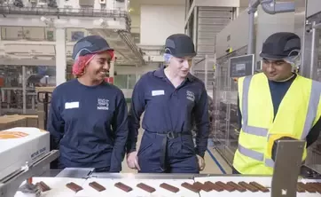 Three apprentices wearing workplace safety equipment standing beside a production line in a food manufacturing facility.