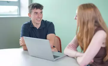 Two people seated at a table discussing work with a laptop open between them.