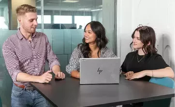 Three people seated at a meeting table discussing work while using a laptop in an office environment.