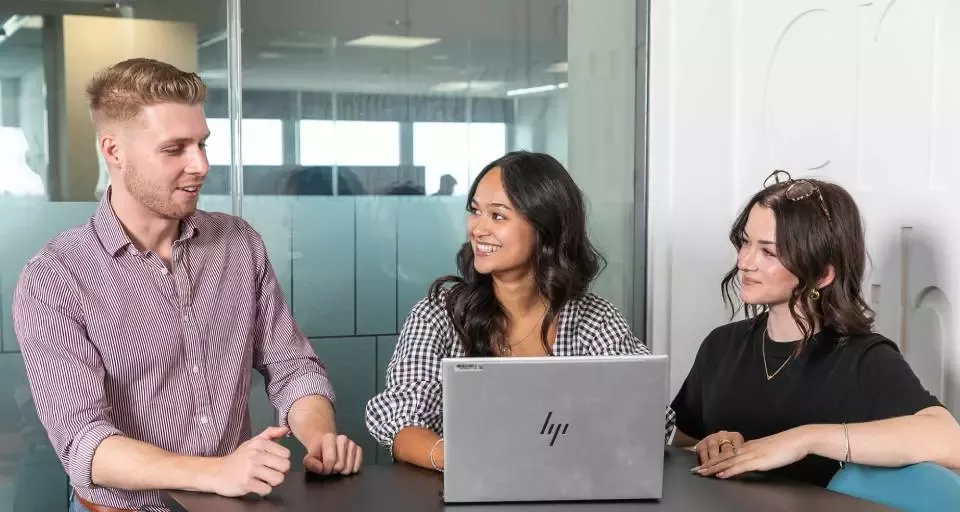 Three people seated at a meeting table discussing work while using a laptop in an office environment.