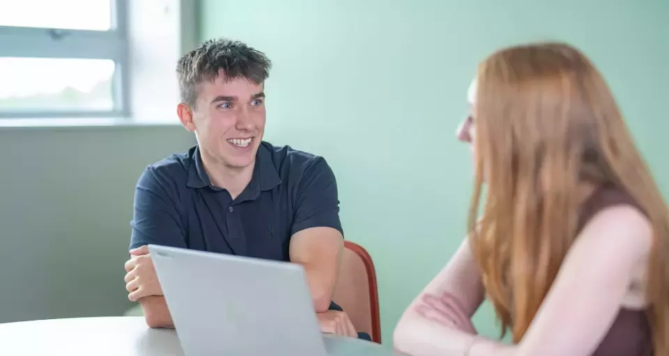 Two people seated at a table discussing work with a laptop open between them.