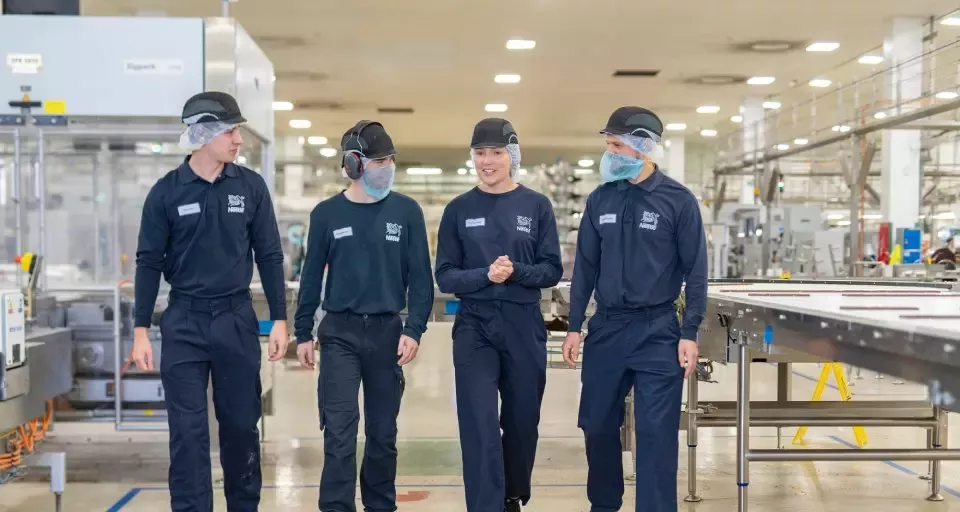  Four people in factory workwear walking through a manufacturing facility alongside a production line.
