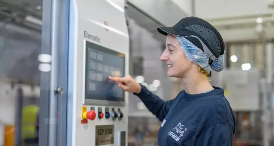 Apprentice operating a touchscreen control panel on manufacturing equipment in a food production facility while wearing workplace safety gear.