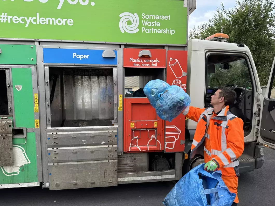Waste collection worker putting blue bin bag into plastics section on truck. 