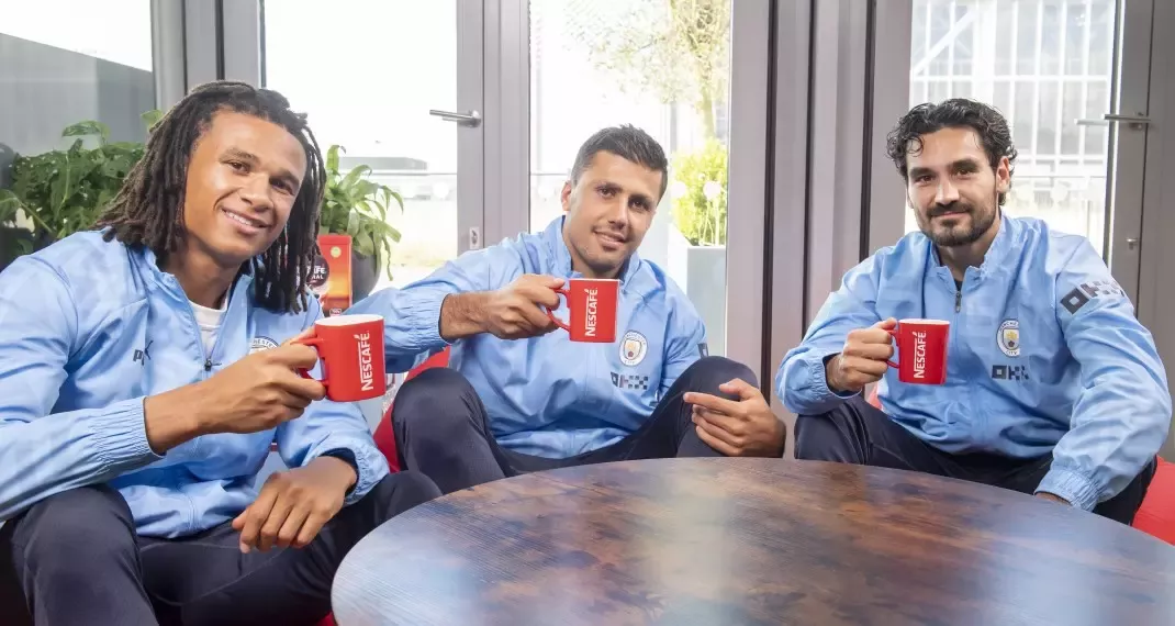 Manchester City football players Ilkay Gundogan, Nathan Ake and Rodri sit down with a cup of coffee in red Nescafé mugs