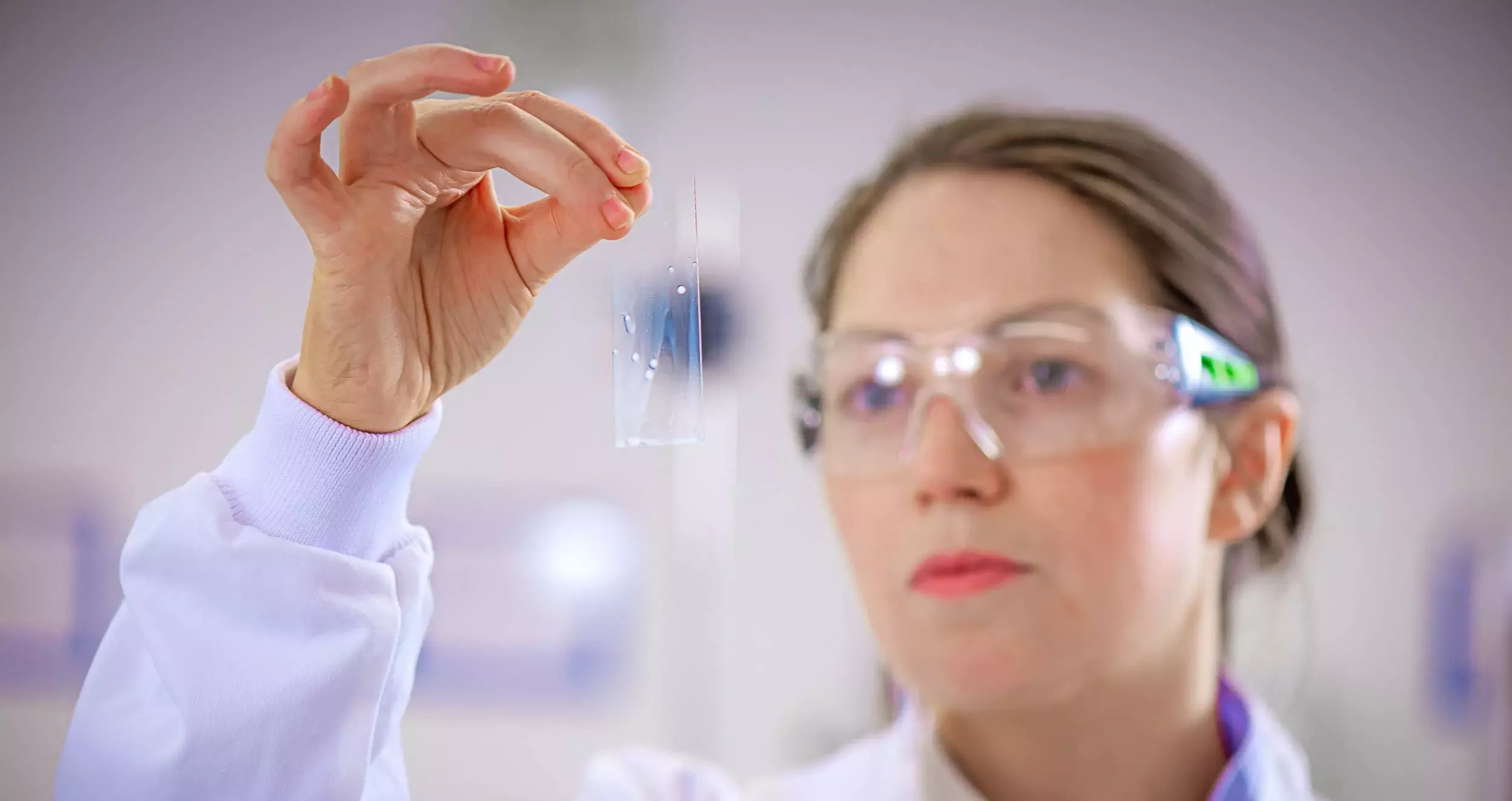 Woman in a lab coat and protective goggles holding a microscope slide.