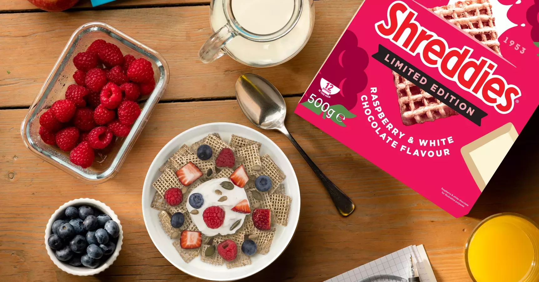 On a table: a bowl of blueberries, a bowl of raspberries, a jug of milk, a spoon, a bowl of Shreddies, and a box of Shreddies