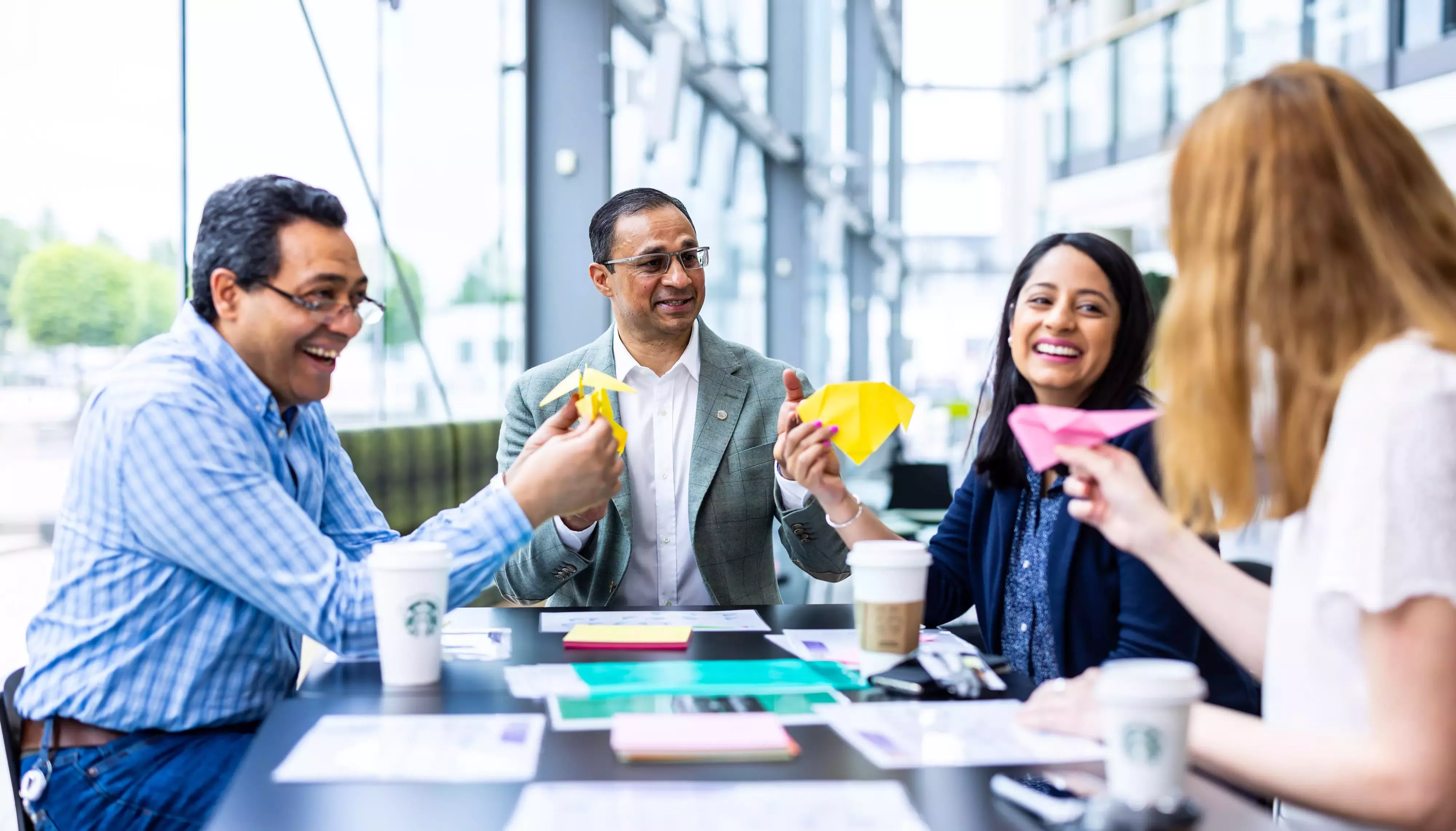 Four individuals gathered around a table holding paper aeroplanes in a workplace setting.