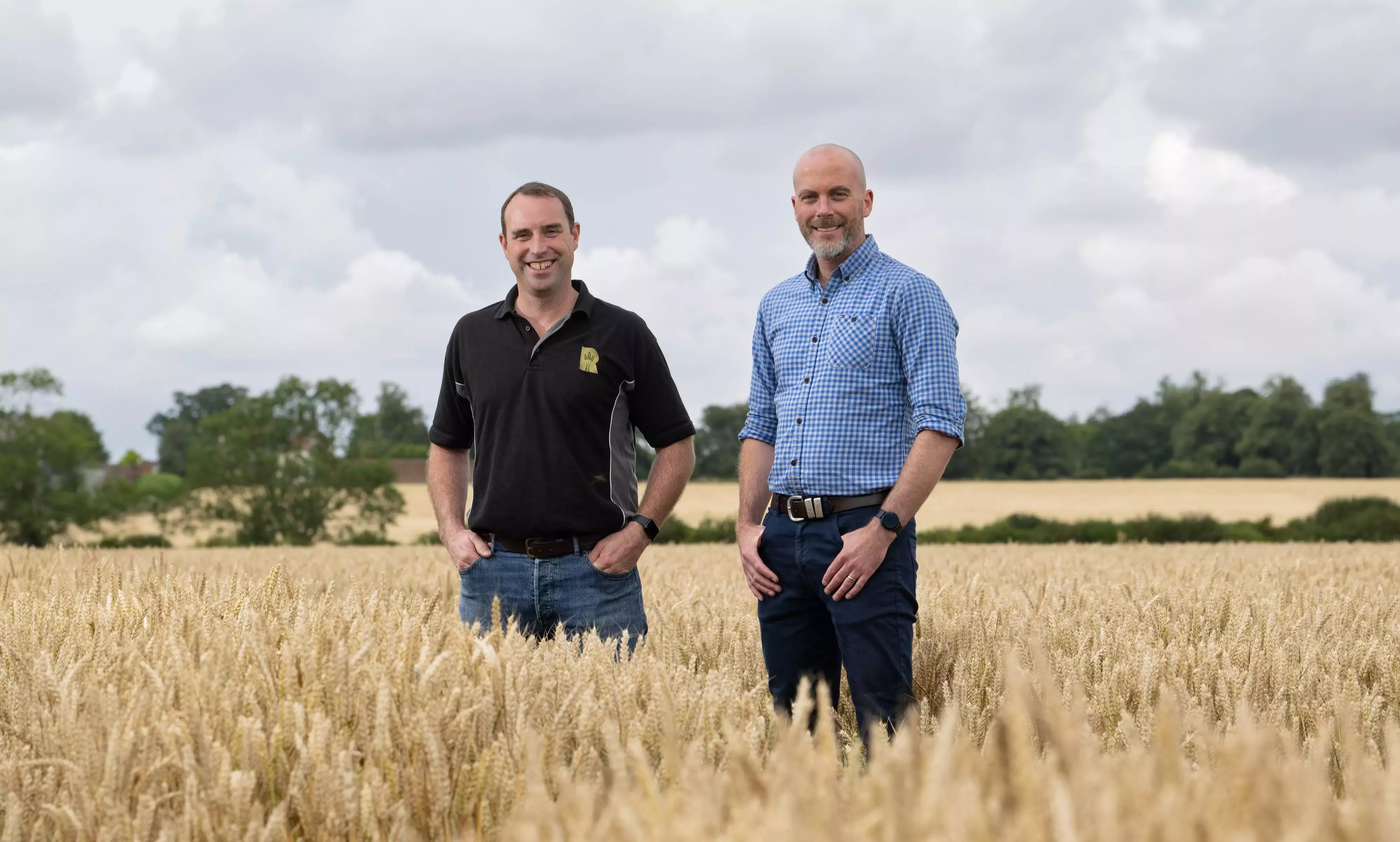 Two men standing next to another in a wheat field with a cloudy sky in the background.